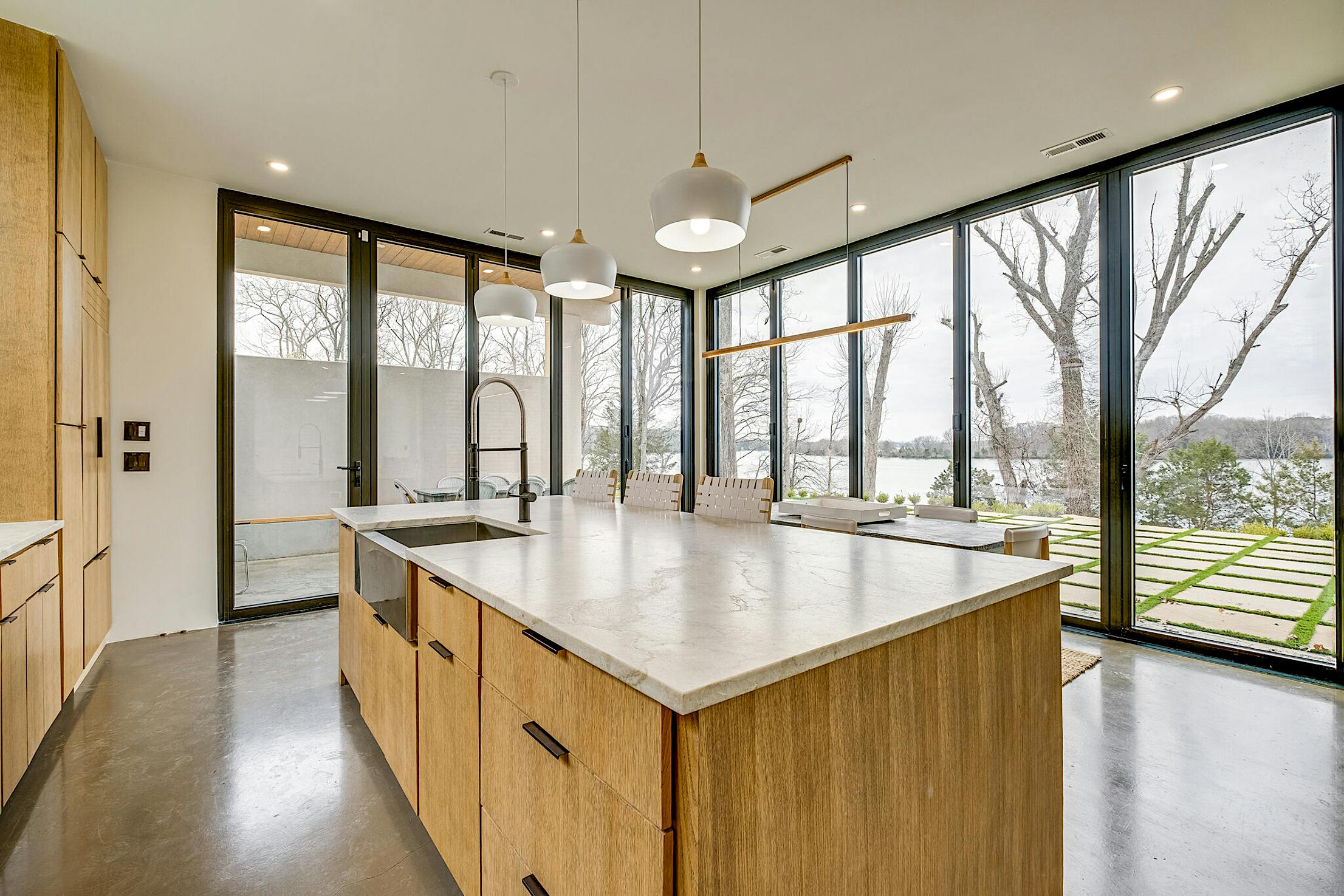 A modern kitchen featuring a large island with a stone countertop and large opening folding glass walls offering a view of the lake and trees in the background.