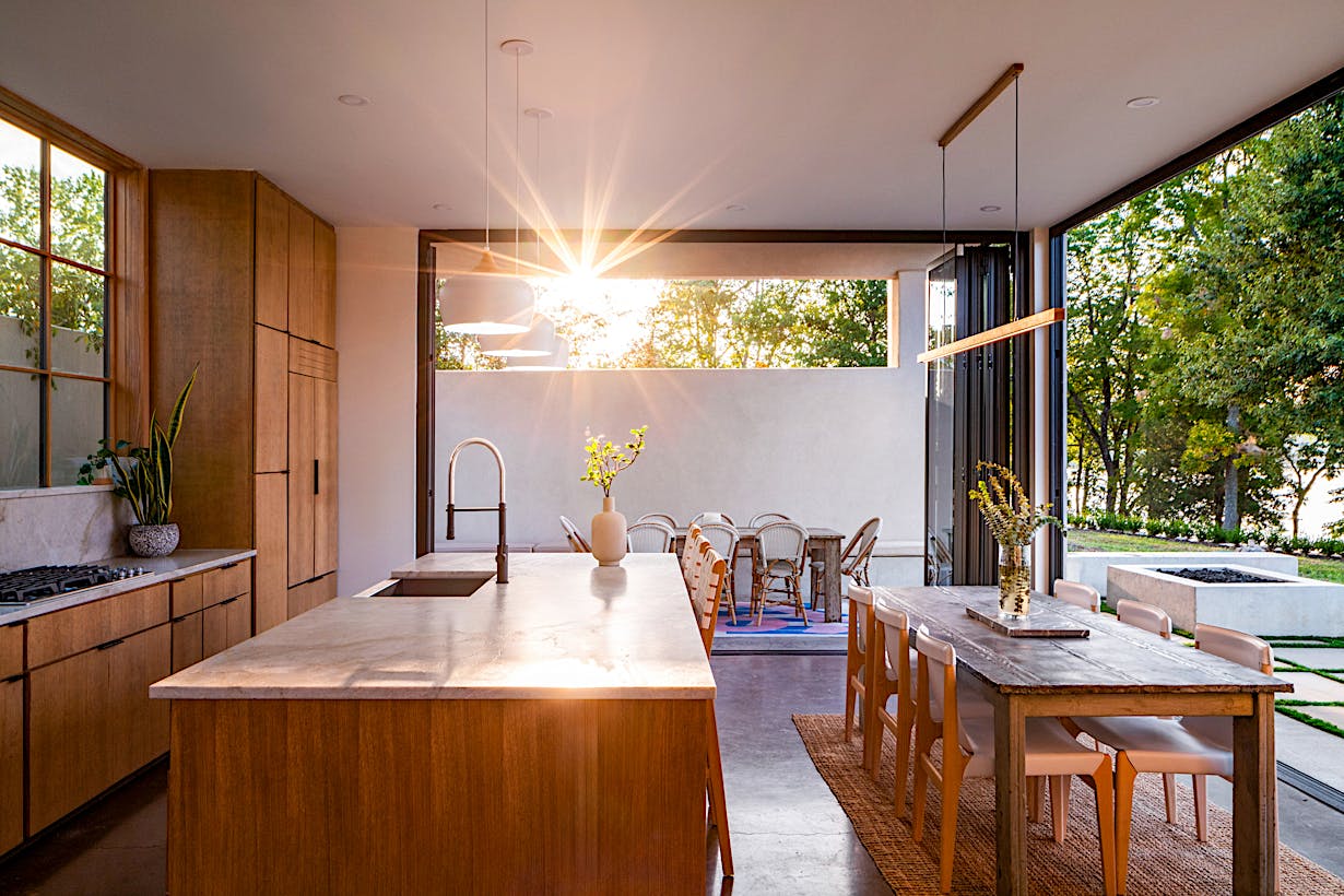 Modern kitchen and dining area with wooden cabinetry and a large island, opening to an outdoor patio. Sunlight streams through the open accordion doors, illuminating the space.