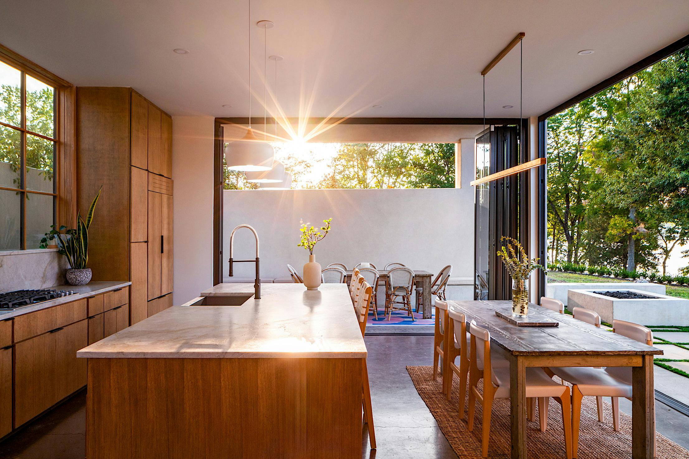 Modern kitchen and dining area with wooden cabinetry and a large island, opening to an outdoor patio. Sunlight streams through the open accordion doors, illuminating the space.