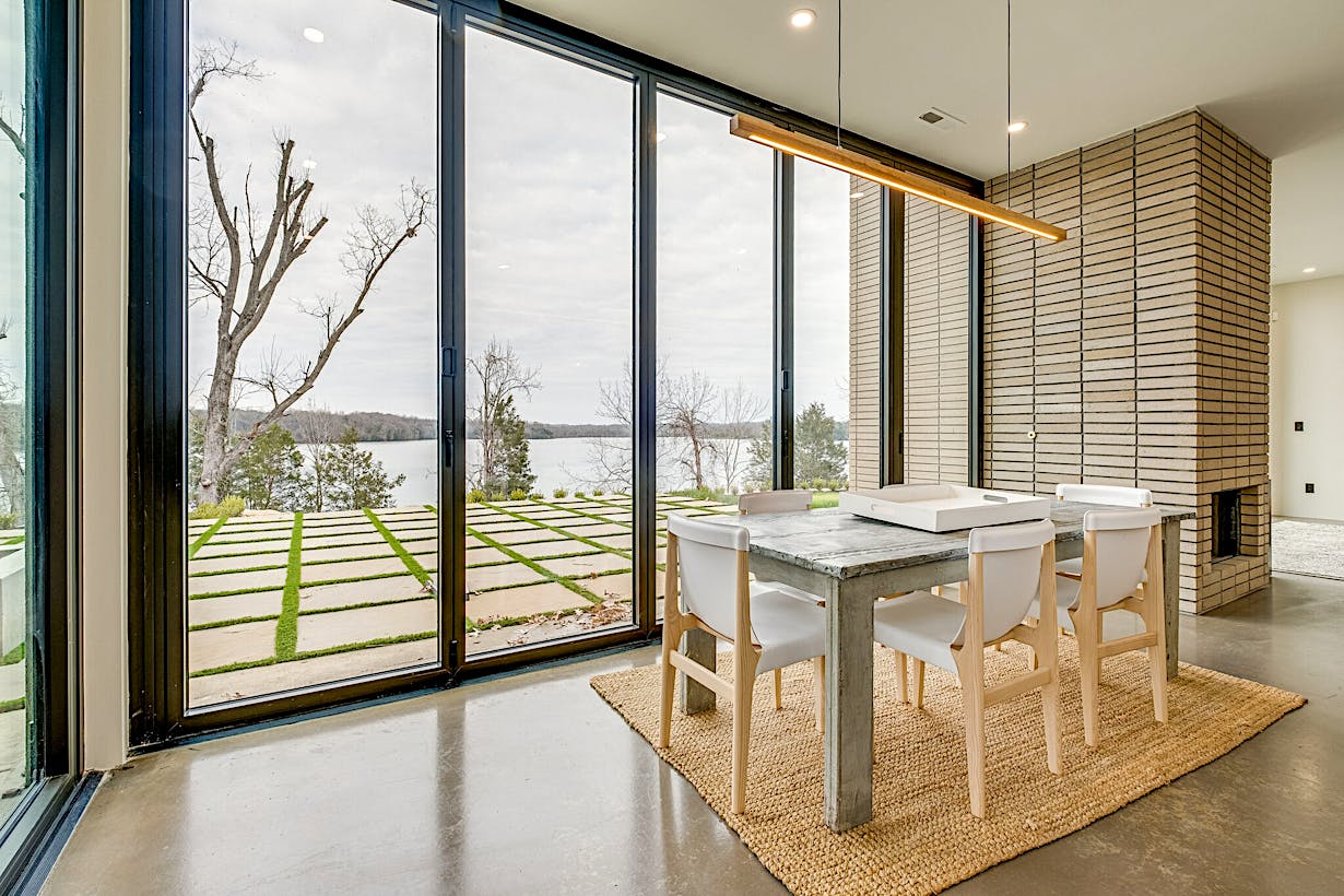 A modern dining area adjacent to floor-to-ceiling glass doors that offer a stunning view of the lake and trees.