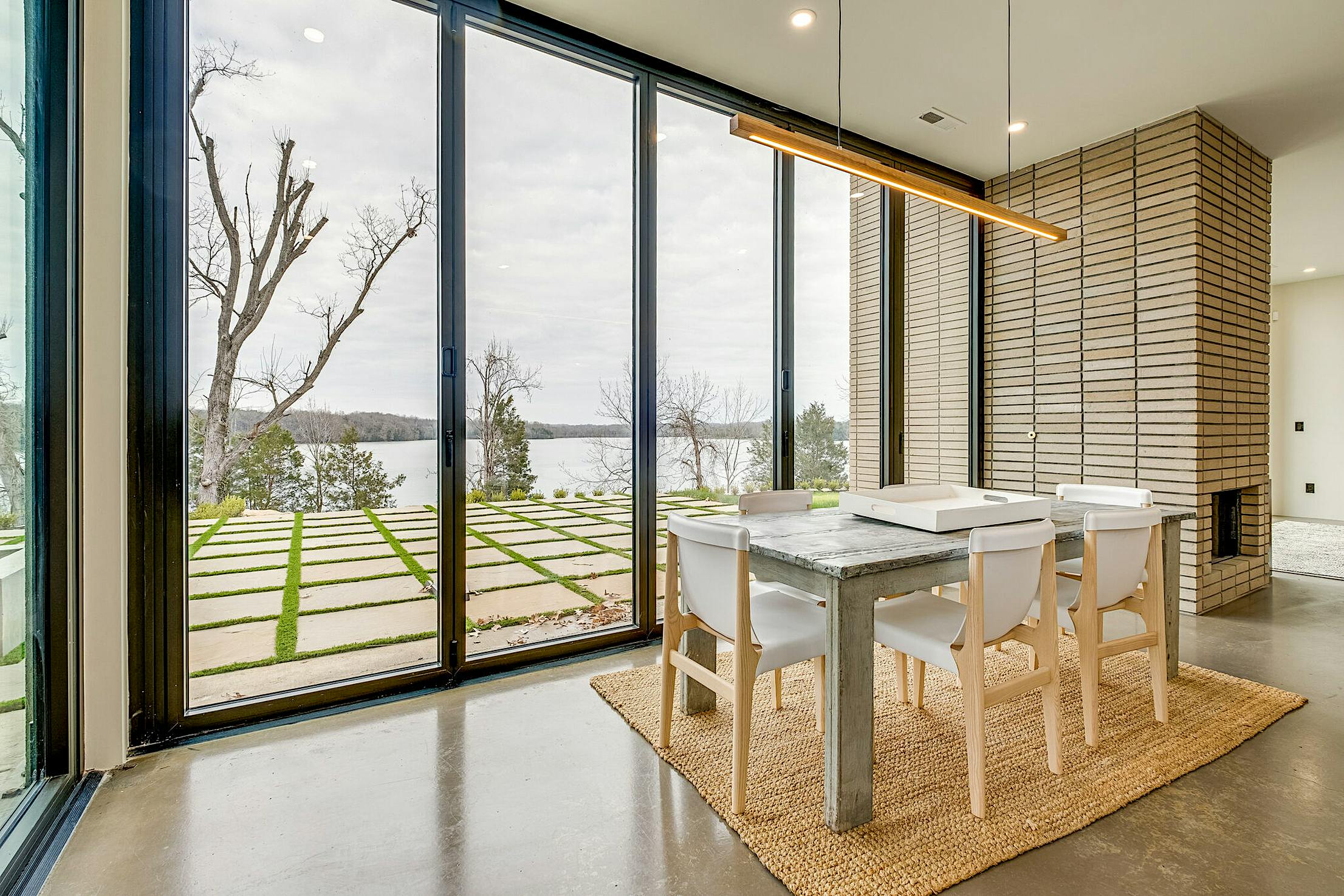 A modern dining area adjacent to floor-to-ceiling glass doors that offer a stunning view of the lake and trees.