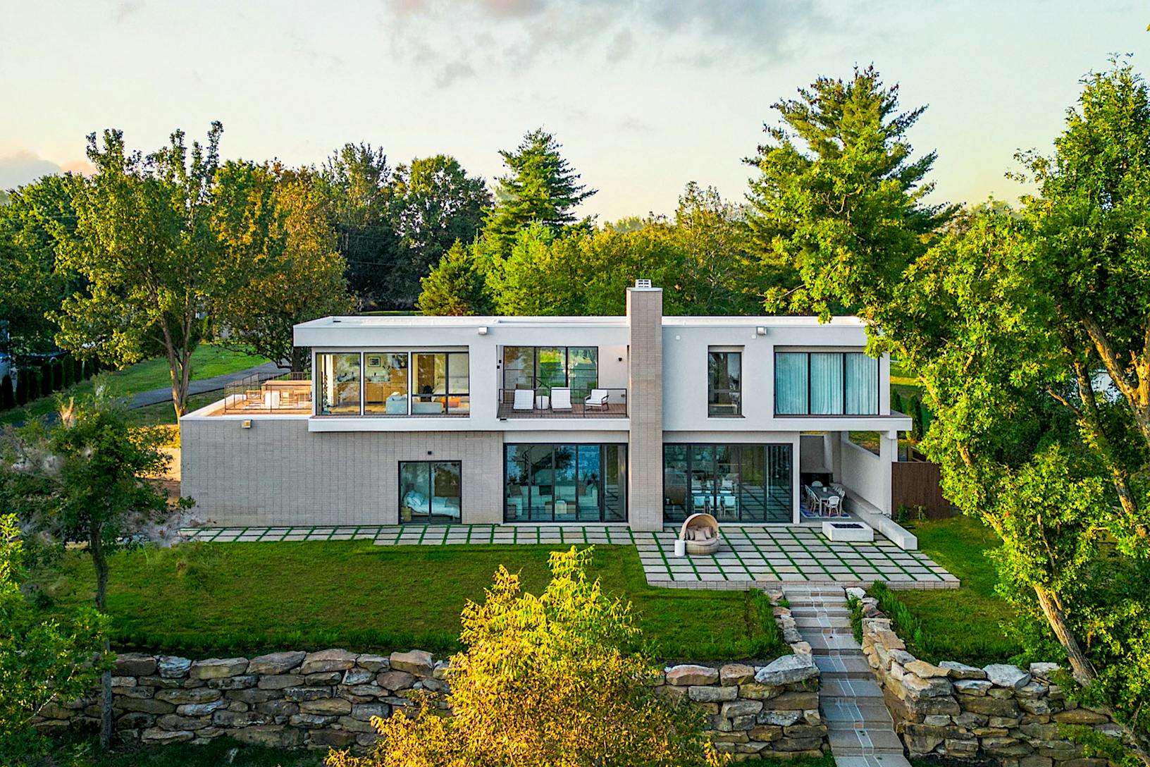 A modern two-story house with large glass wall panels, surrounded by trees and greenery, featuring a spacious patio.