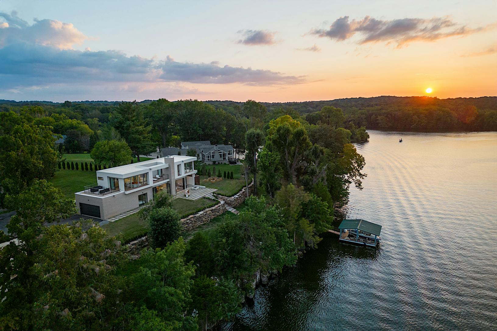 A modern house with large windows stands on the edge of a wooded area overlooking a lake at sunset.