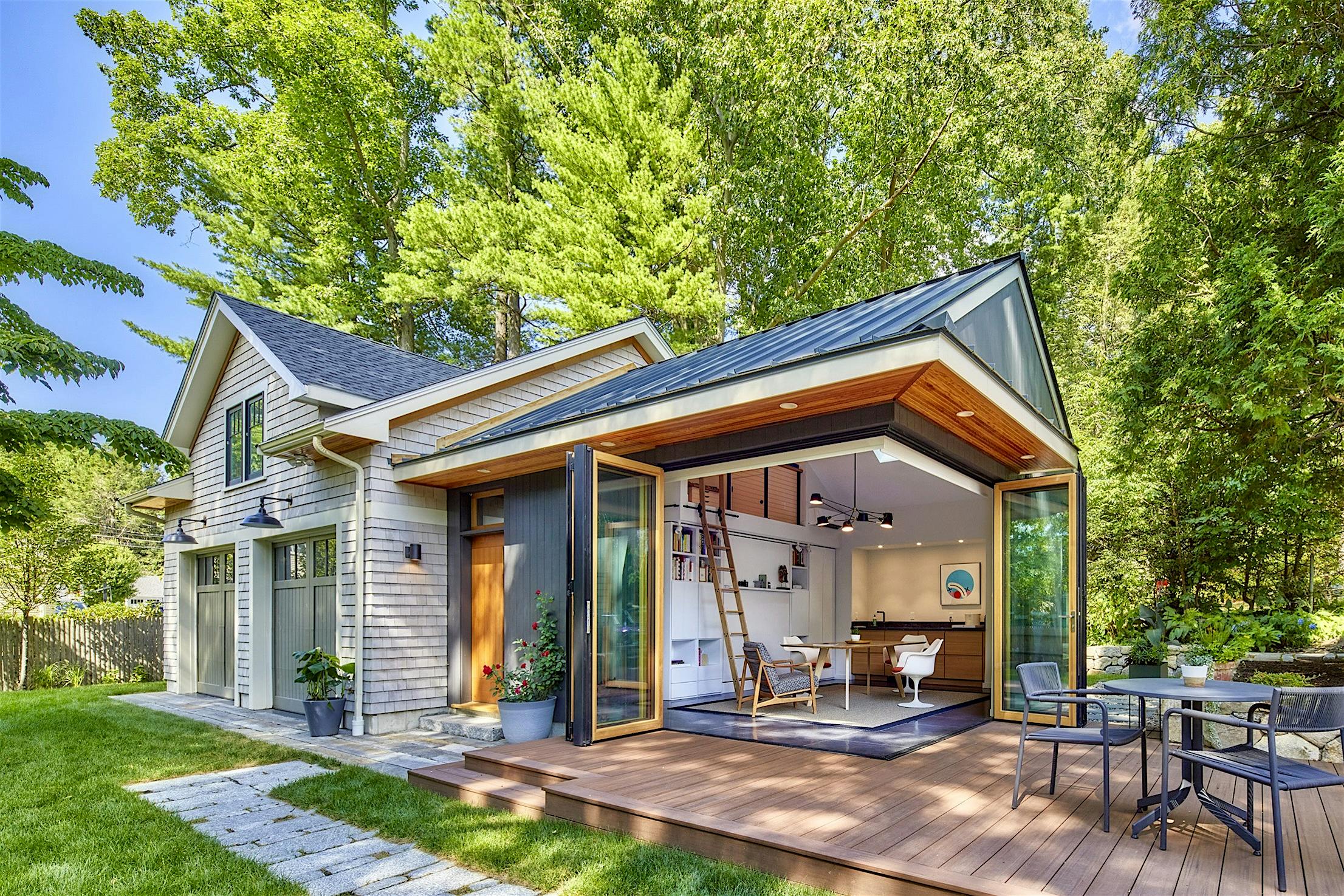 A modern house features cornerless bi-folding doors that open to reveal an interior office space, surrounded by greenery and outdoor seating on a wooden deck.