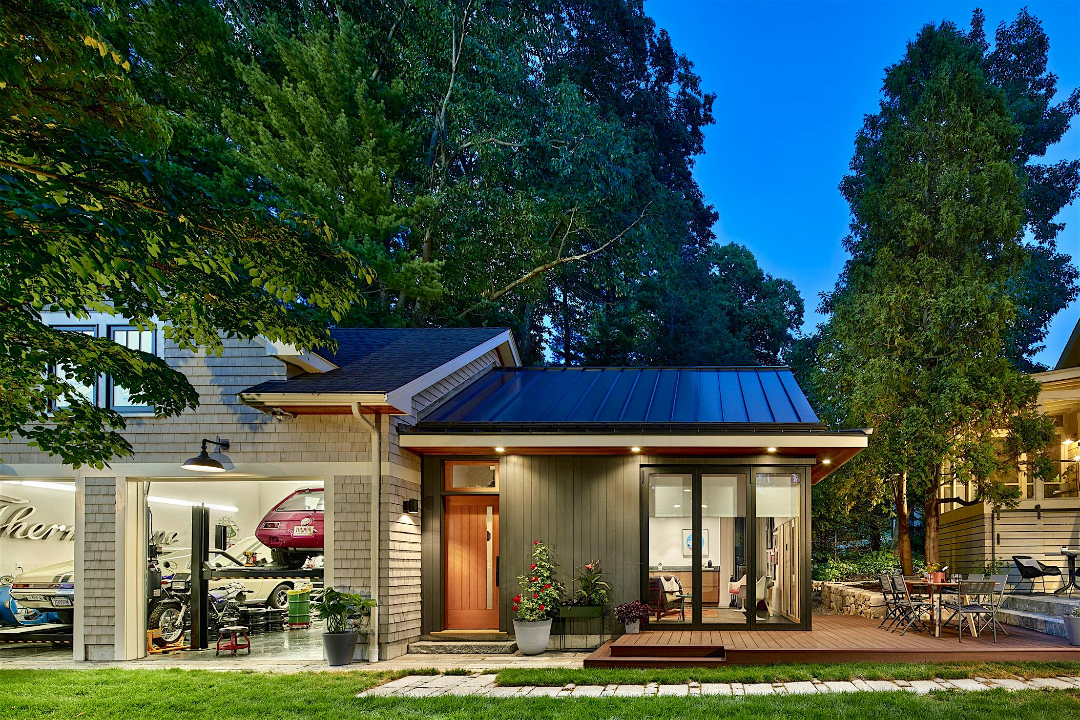 A modern house at twilight with glass bifold doors leading to a garage showcasing cars on the left, large windows, an outdoor seating area, and surrounded by trees.