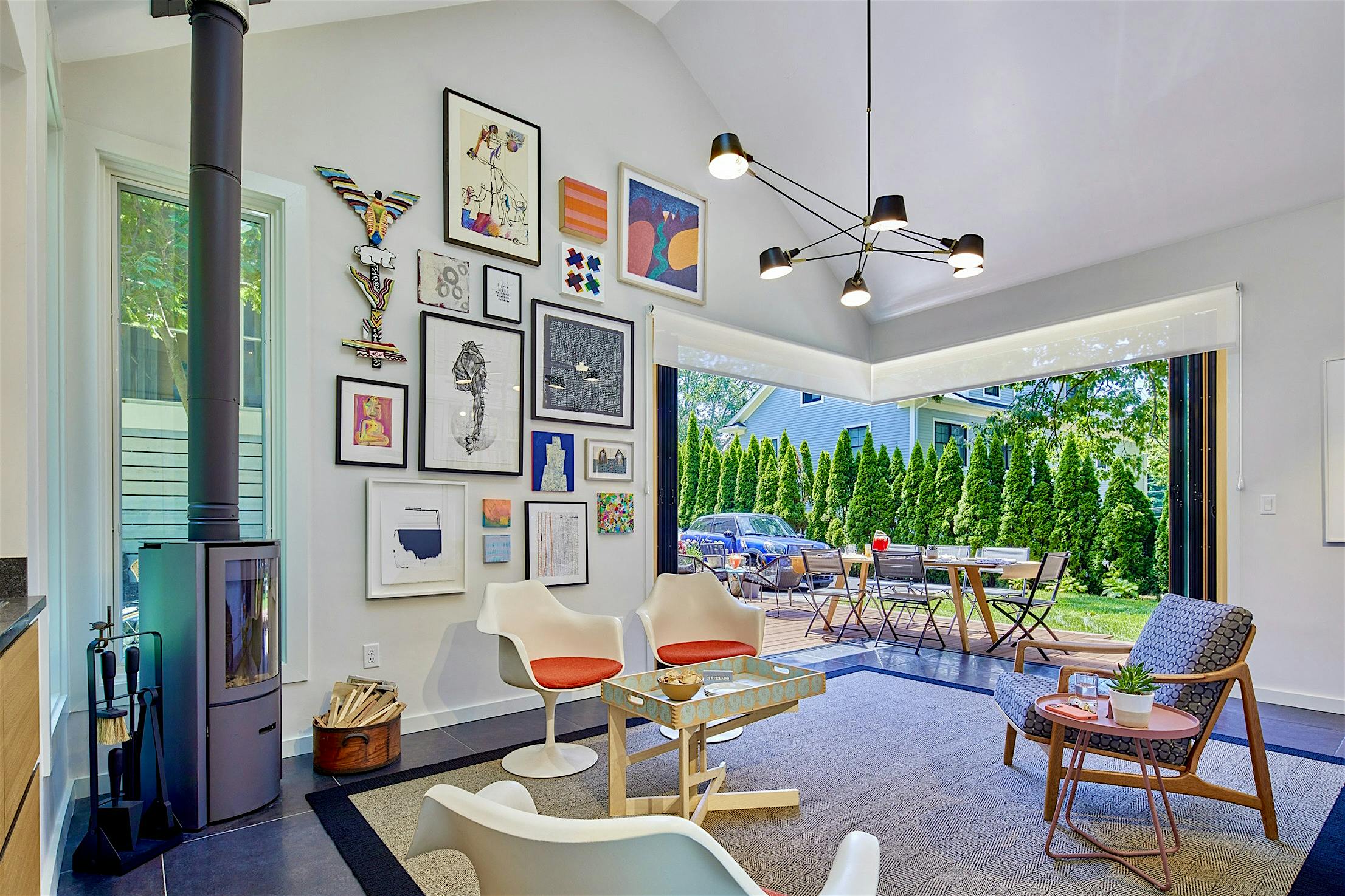 A modern living room with a high ceiling, artwork on the walls, a wood-burning stove, minimalist furniture, and large bifold glass doors opening to a patio with outdoor seating and greenery.