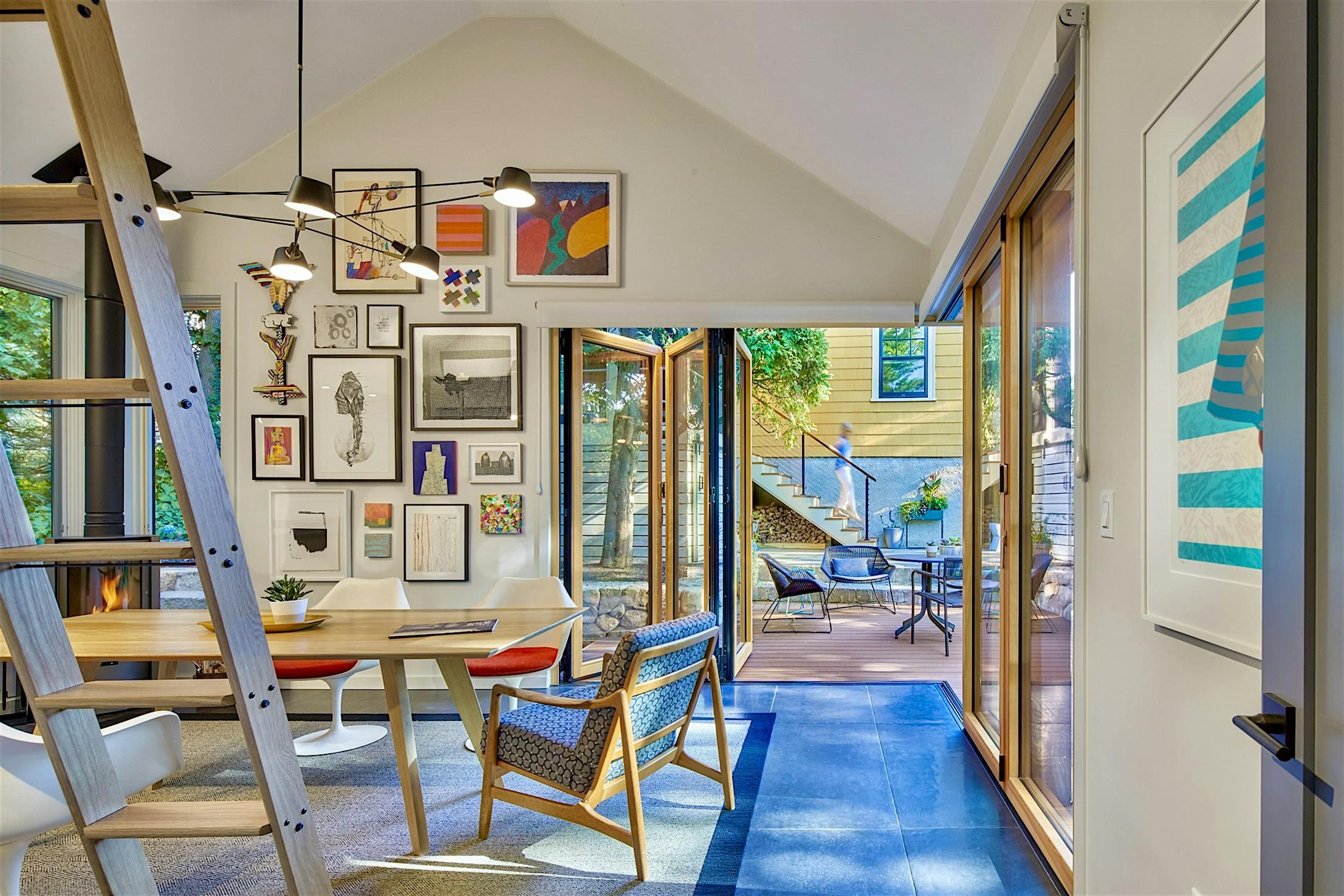 A modern art studio with a wooden table and chairs, a wall displaying various framed artworks, large windows, glass bifold doors leading to an outdoor deck, and a ladder leading to a loft space.