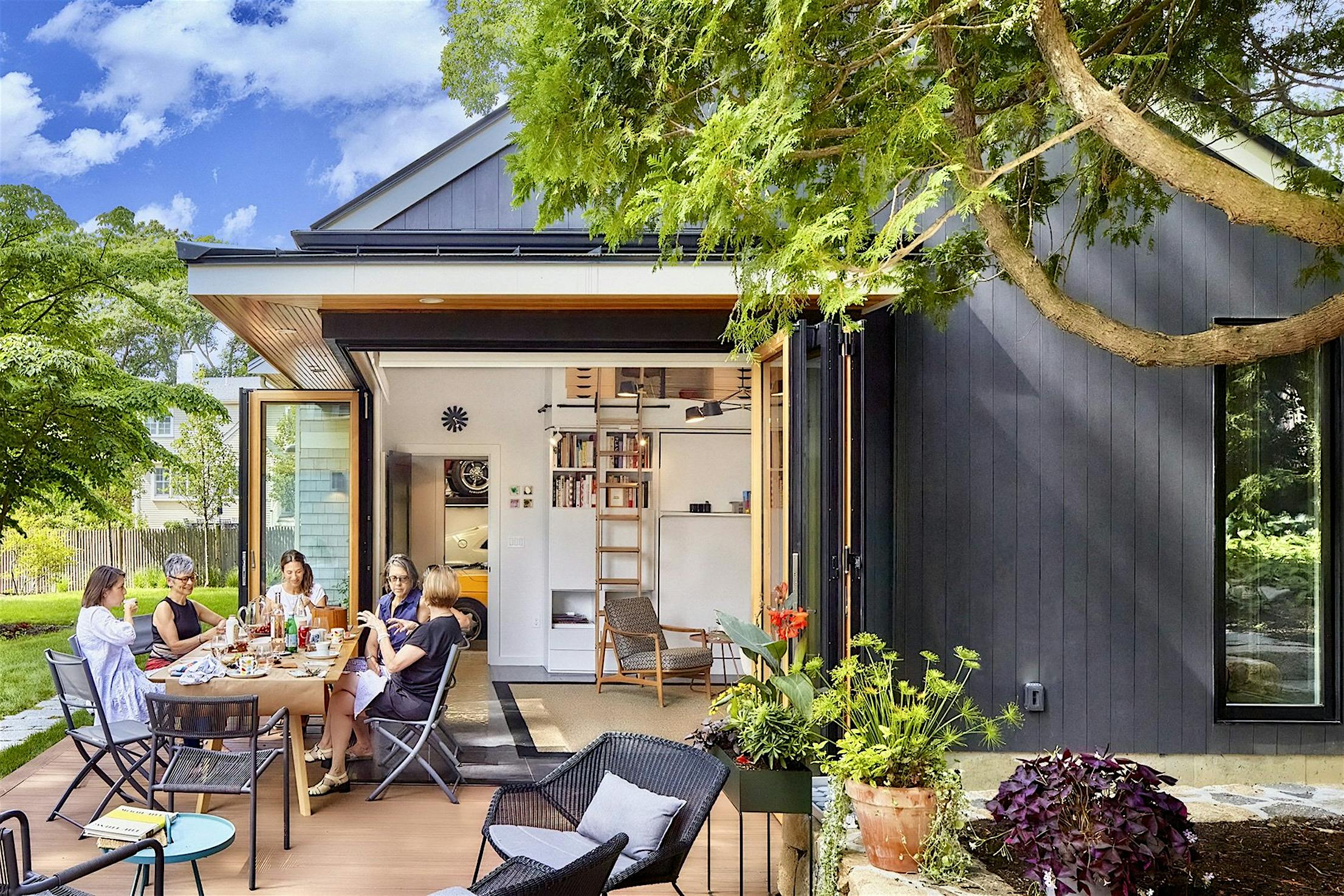 Four people are sitting around a table on a patio next to a modern grey ADU, engaged in conversation and enjoying food. The bifold glass doors are open, revealing an interior with shelves and a ladder.