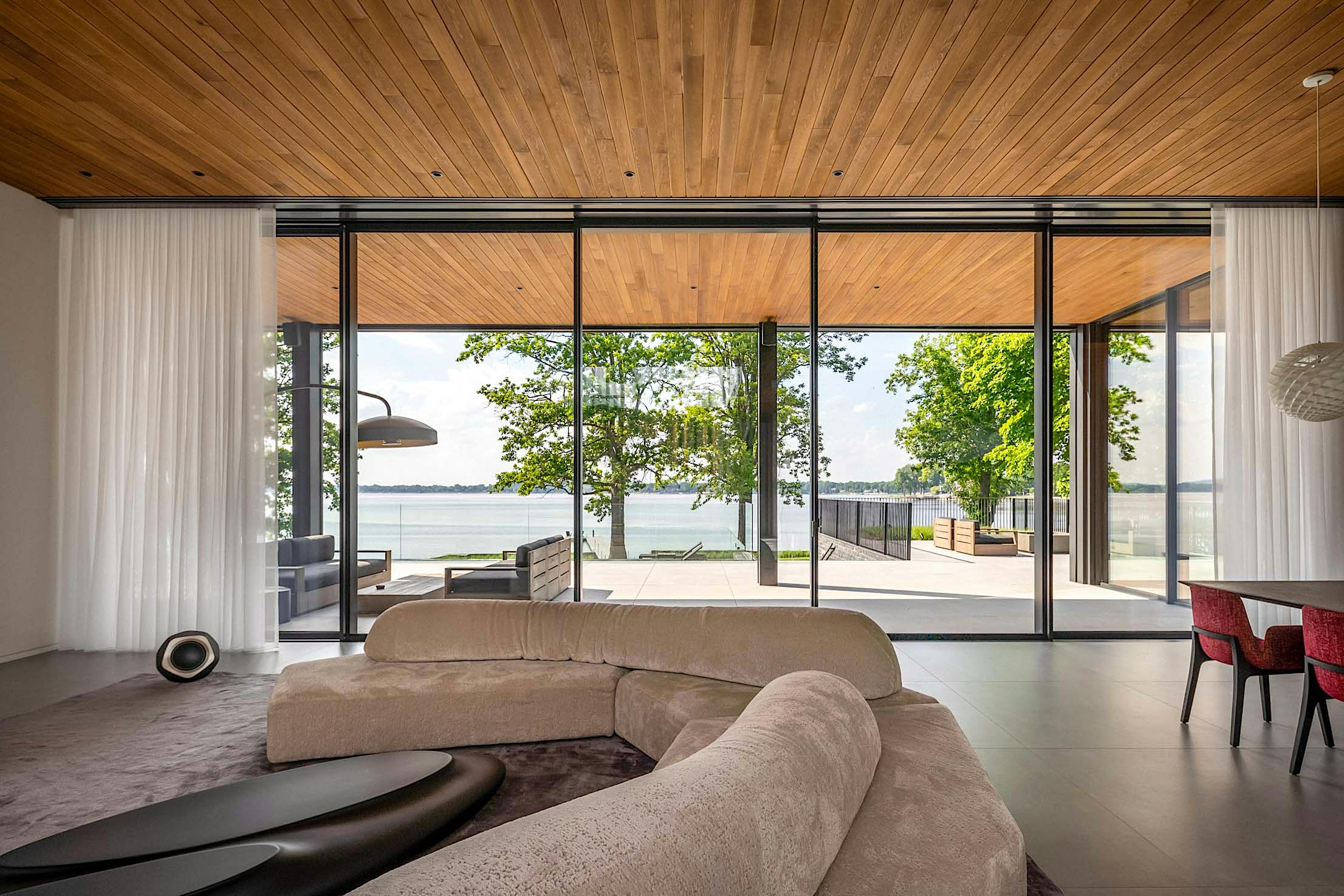 Modern living room with large sliding glass doors, offering a view of a lake and trees. Features a curved sofa, wooden ceiling, and outdoor seating area.