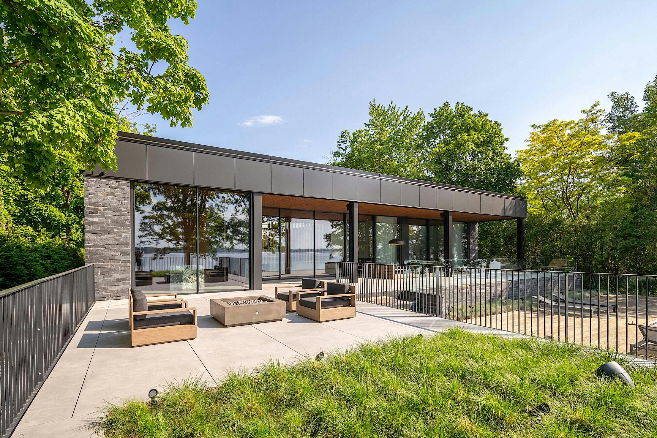 Modern house with large windows and outdoor seating on a patio, surrounded by lush greenery and a view of a body of water in the background.