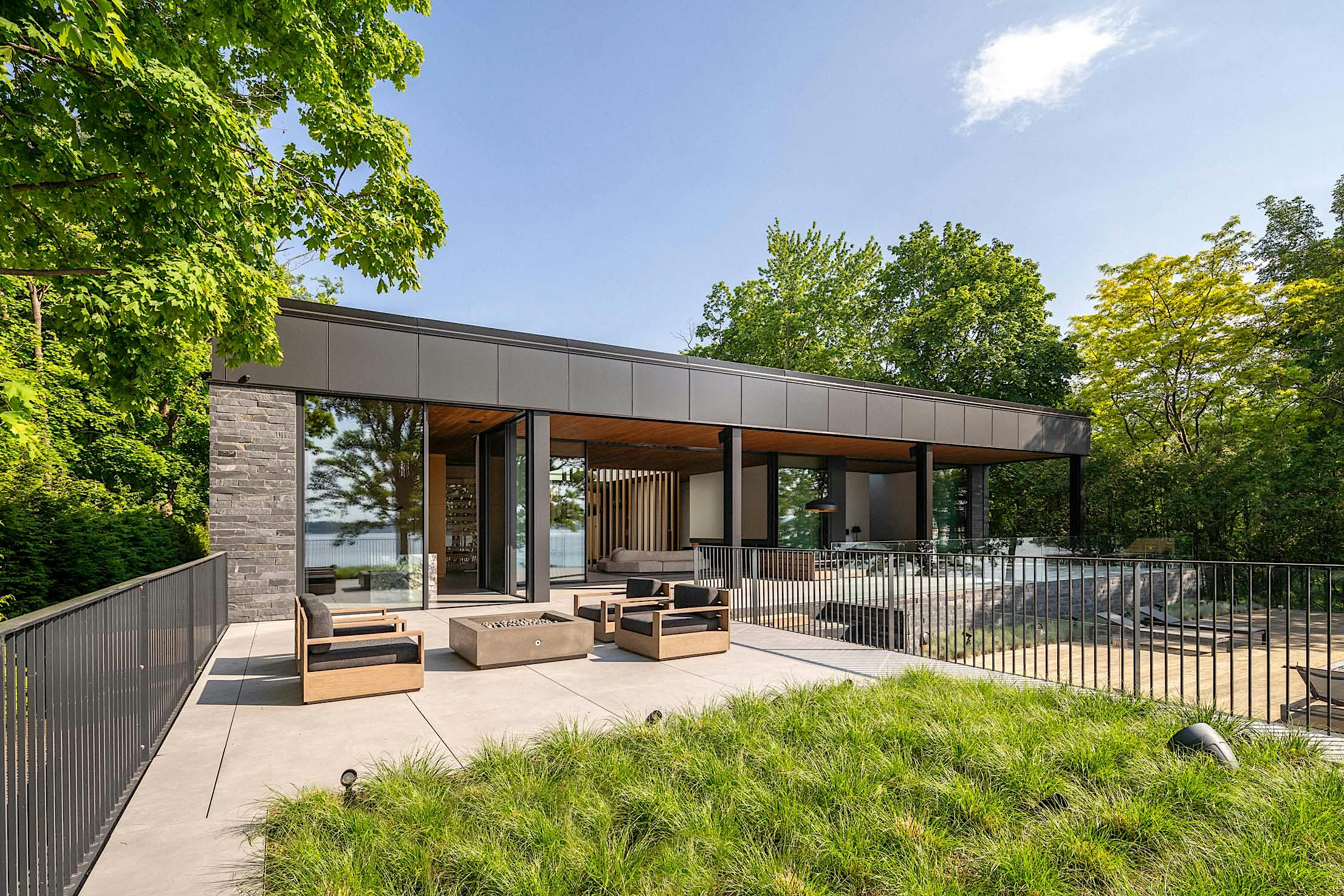 Modern open-air patio with seating area and fire pit, surrounded by greenery and trees, adjacent to a contemporary building with large glass windows under a clear blue sky.