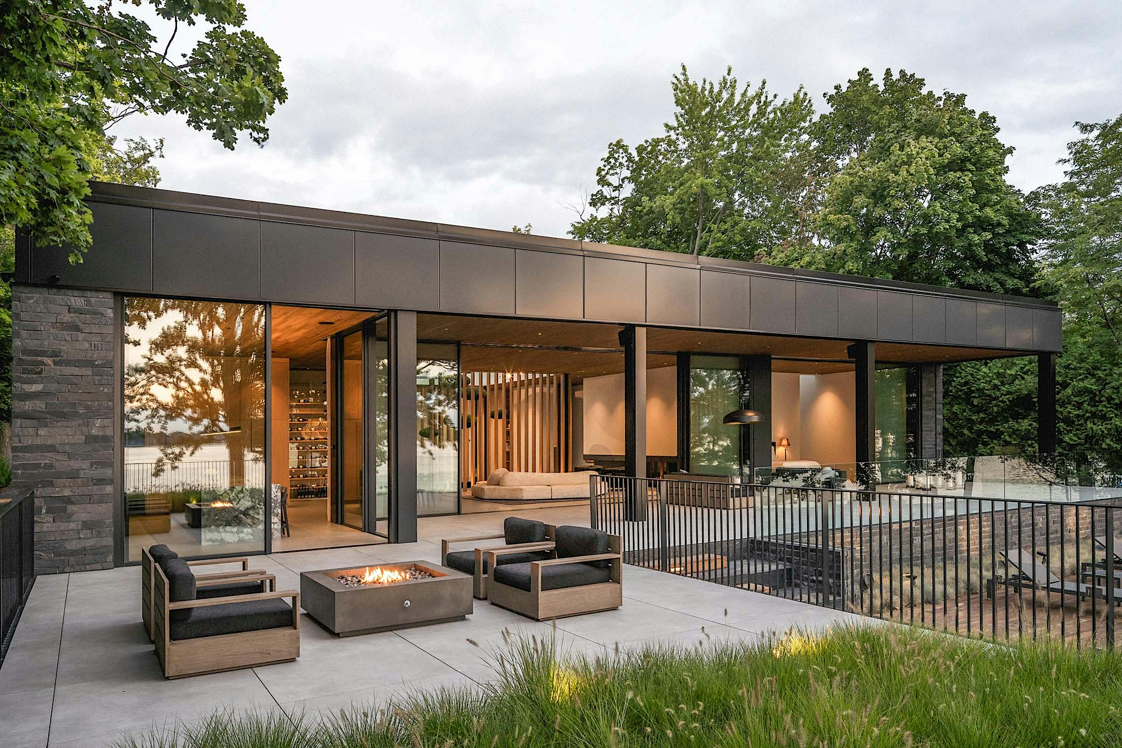 Modern house with large glass doors, a covered patio, and an outdoor fire pit surrounded by chairs, set against a backdrop of trees and greenery.