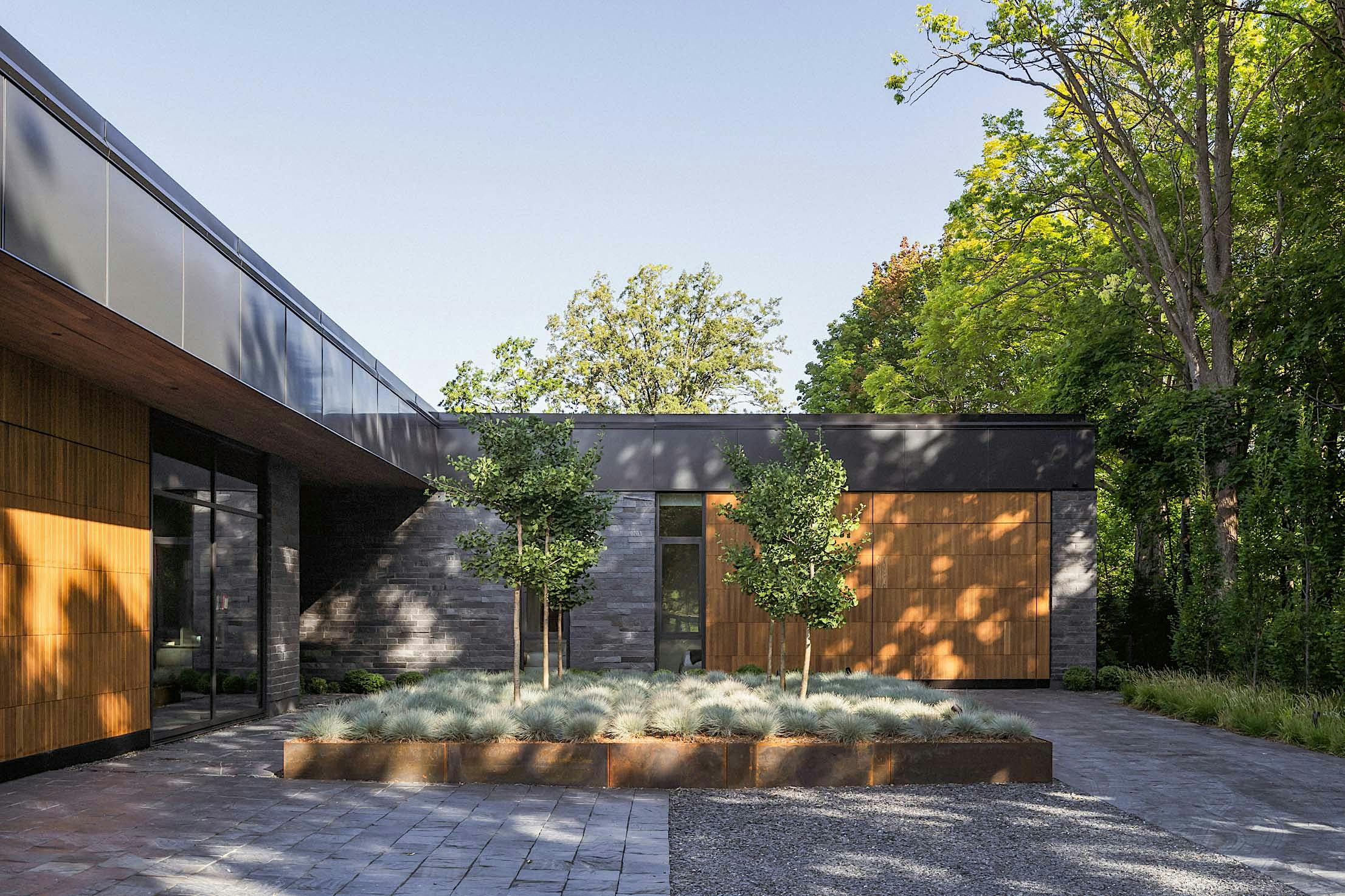 Modern house exterior with wood and stone facade, large sliding glass doors, and a landscaped courtyard featuring two small trees and shrubs.