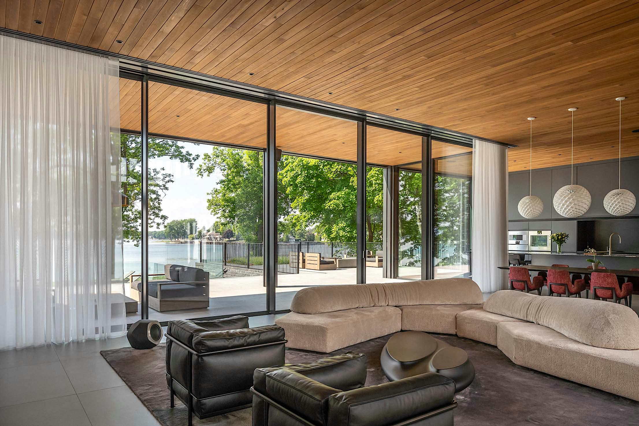 Modern living room with large sliding glass doors, brown couches, and two black chairs. Overlooks a patio and lake with trees.