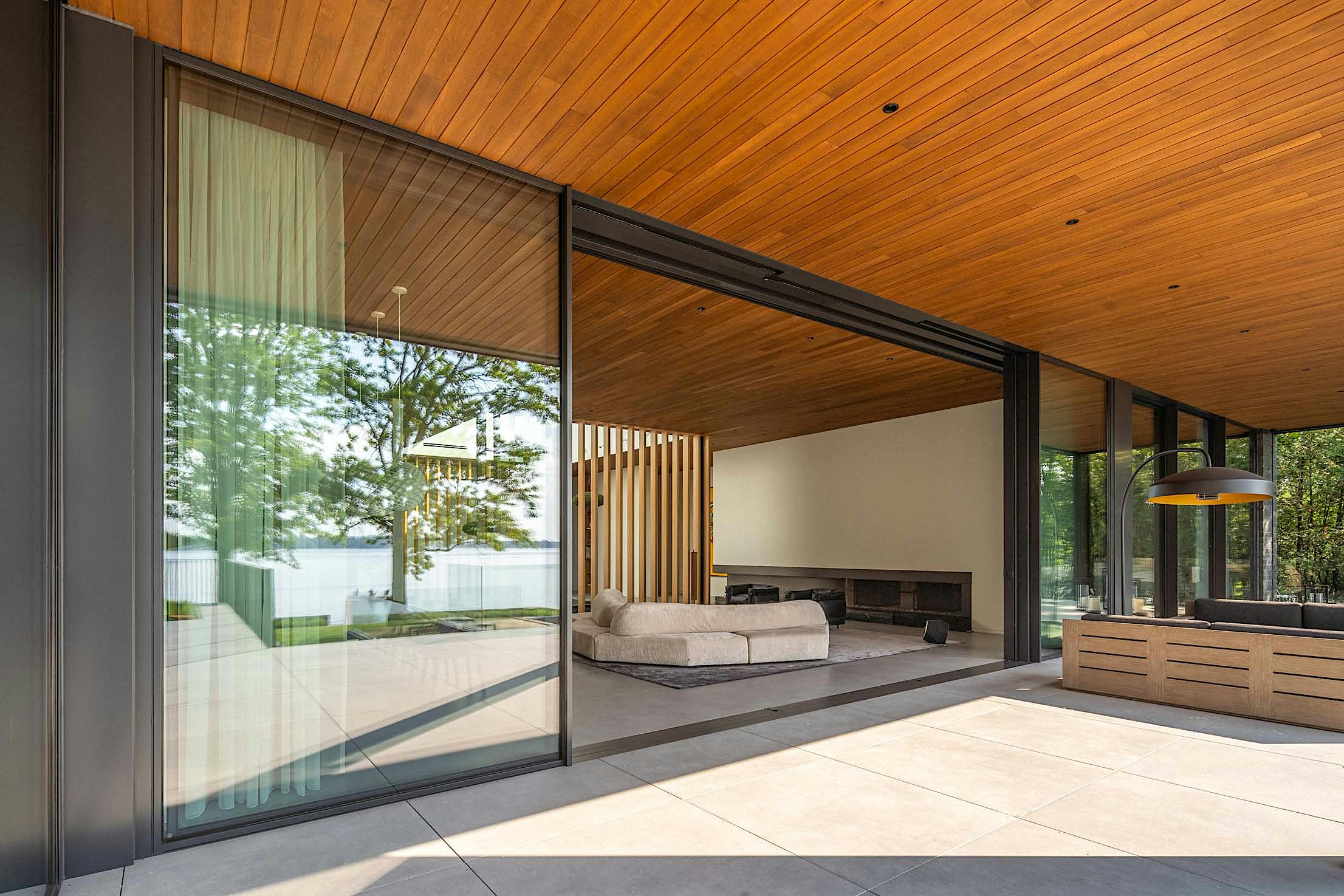 Modern open-concept living space with large glass doors, wooden ceiling, and a view of trees and water.