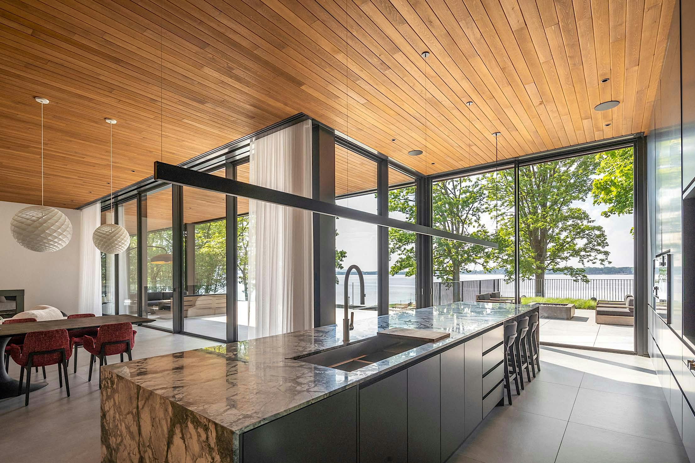 Modern open-concept kitchen and living room with large glass doors, a marble island, black cabinets, and a view of trees and water outside.