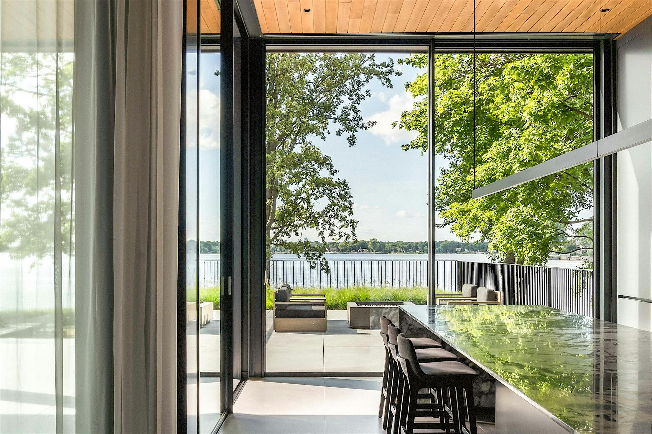 Modern dining area with large glass doors showcasing a scenic view of a patio, trees, and a lake. Minimalist design featuring a long table and chairs.