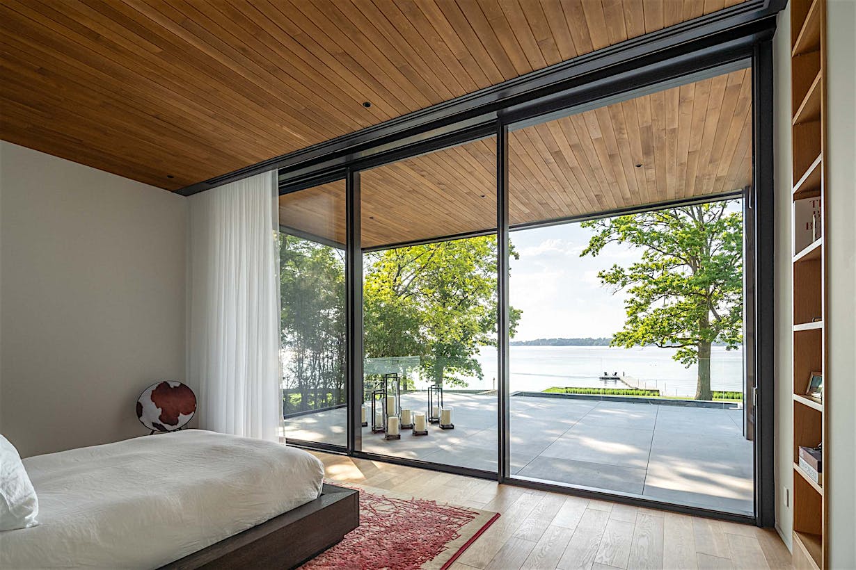 A modern bedroom with large glass windows overlooks a patio and a serene lake view.