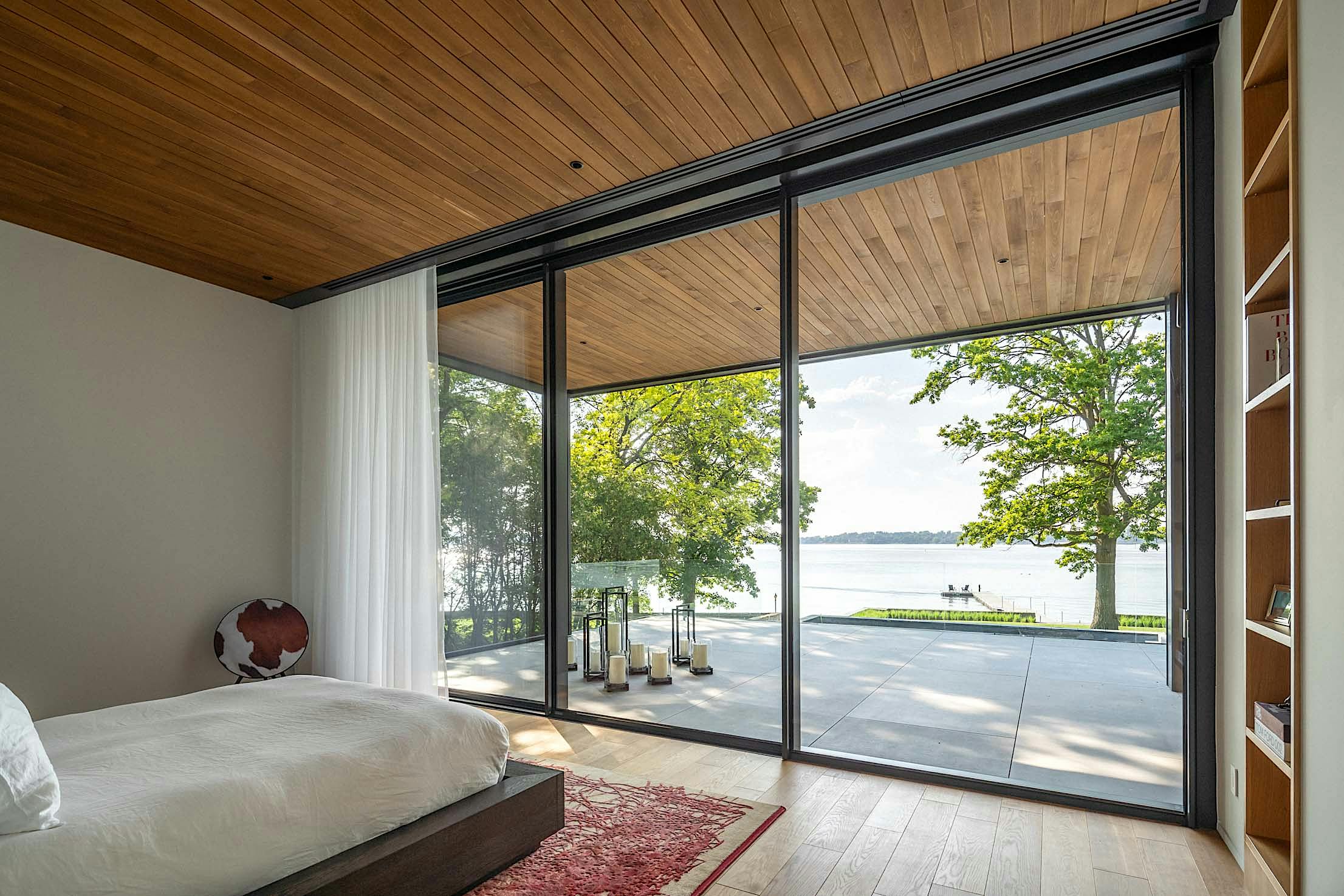 A modern bedroom with large glass windows overlooks a patio and a serene lake view.