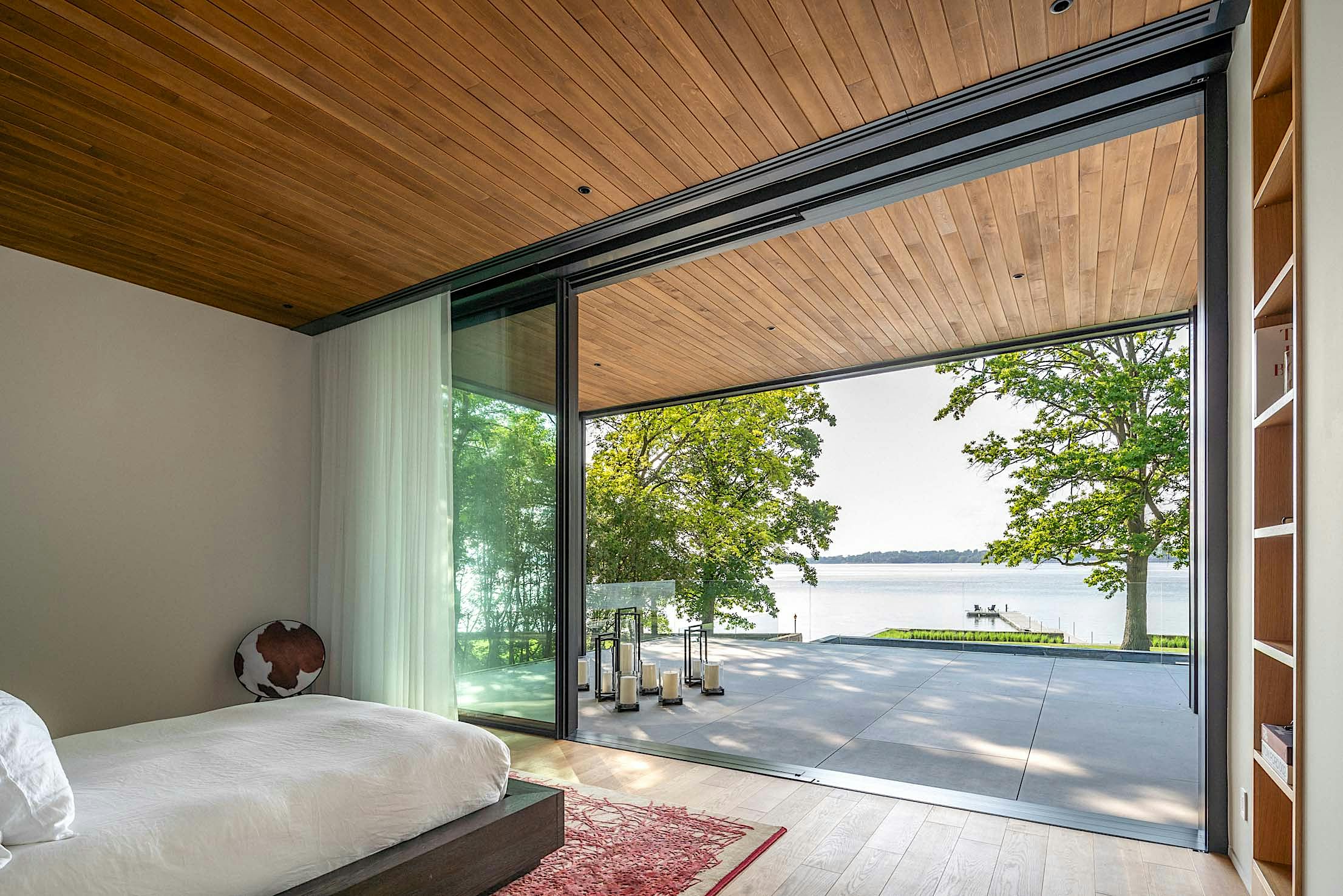 A modern bedroom with a wooden ceiling, large glass doors, and a view of a lake and trees.