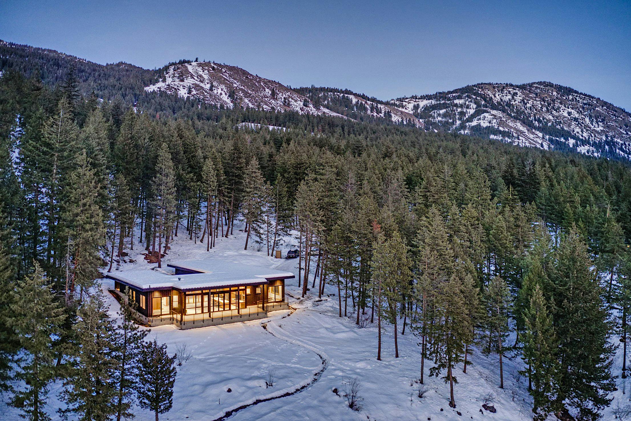 A modern house with accordion glass doors and large windows is surrounded by snow-covered trees and mountains under a clear sky.