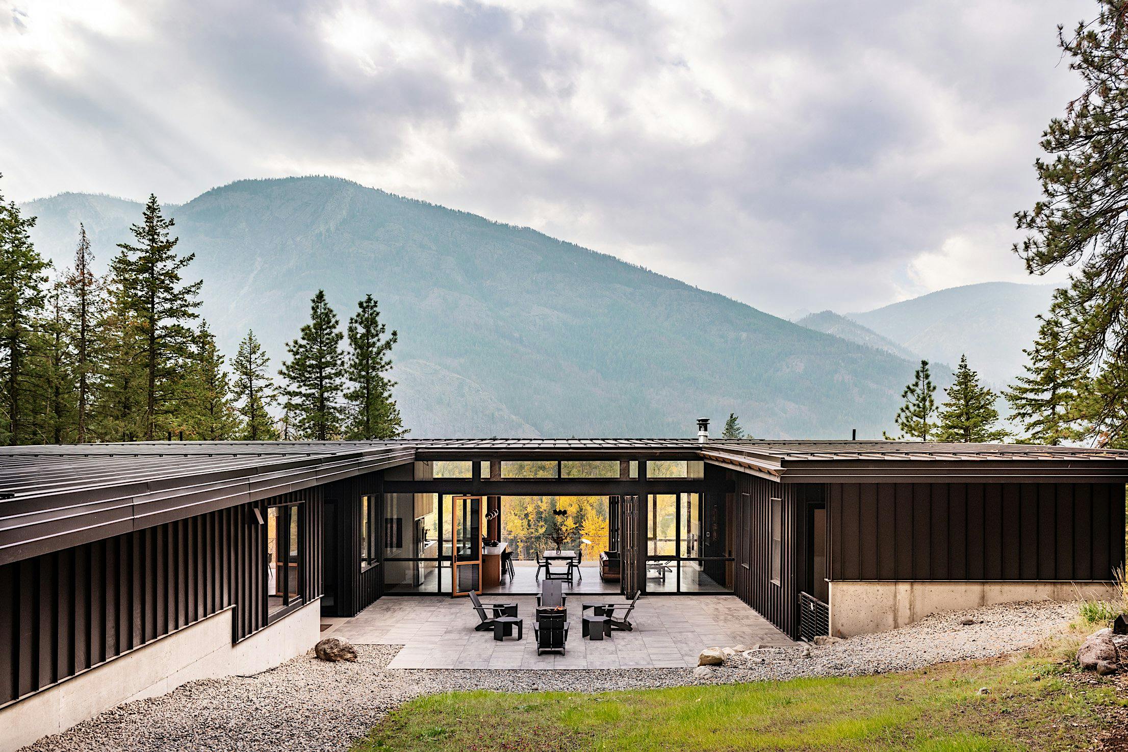 Modern house with folding glass doors leading to a large patio, surrounded by trees and mountains under a cloudy sky.