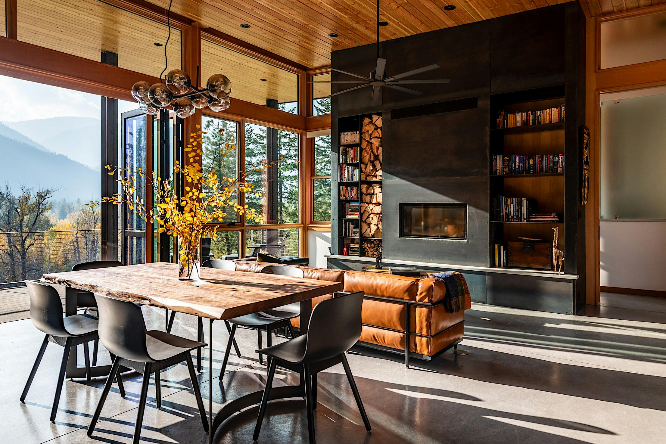 Modern living and dining area with a wooden table, black chairs, fireplace, and large folding doors showcasing a scenic mountain view.