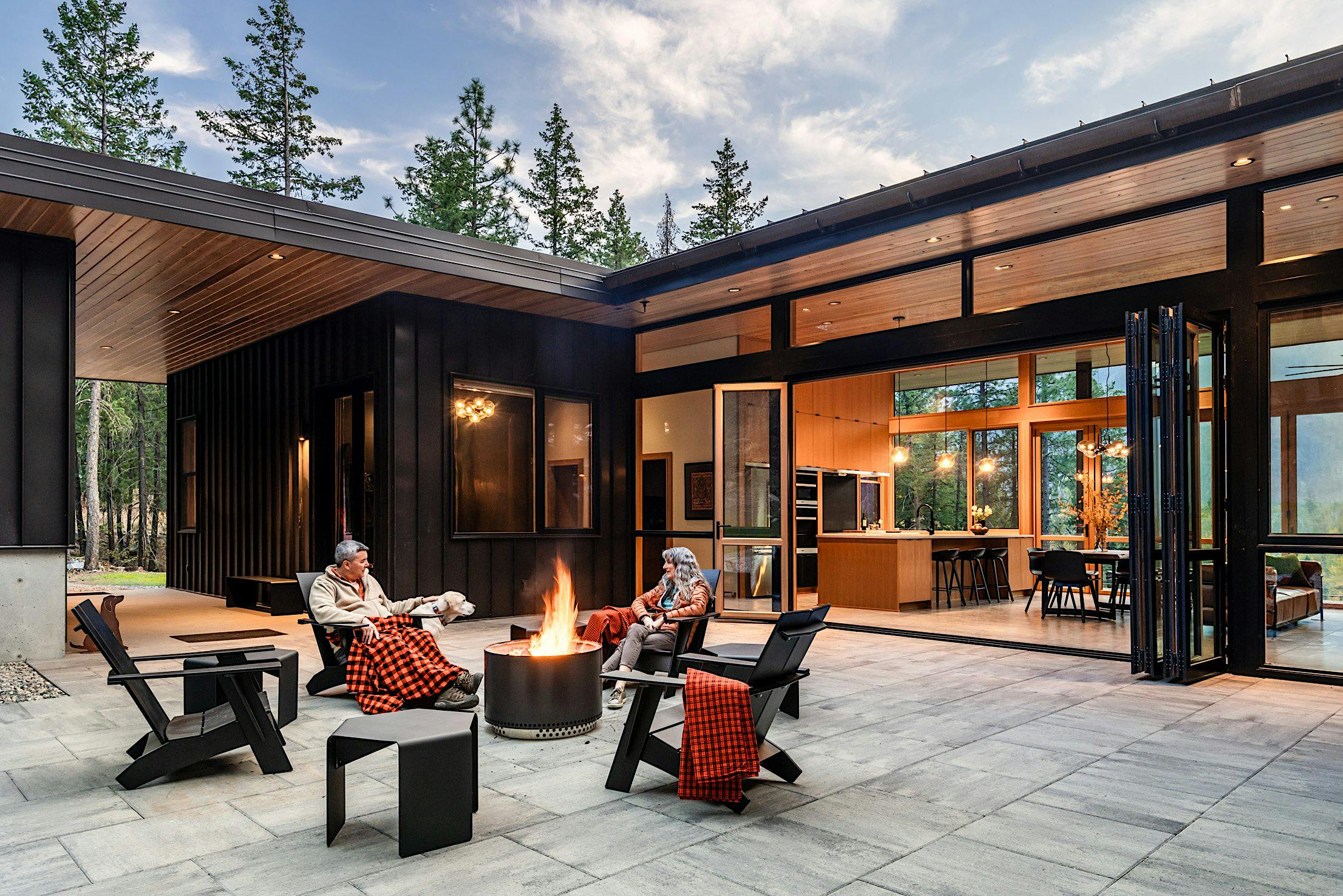 Two people sit by a fire pit on a modern patio, surrounded by tall trees. The house features large accordion glass doors and an open living area visible in the background.