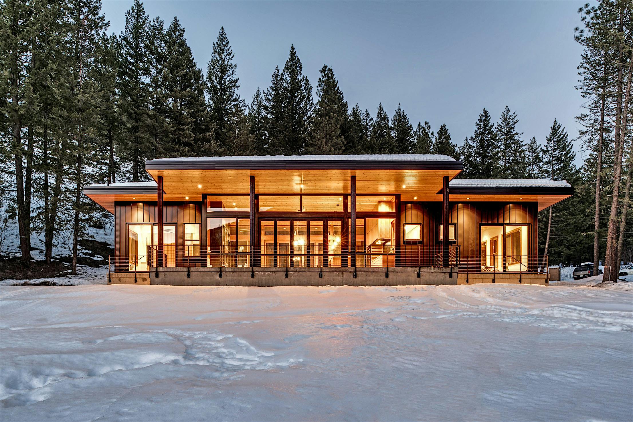 Modern cabin with large windows and warm interior lighting, surrounded by snow and tall pine trees.