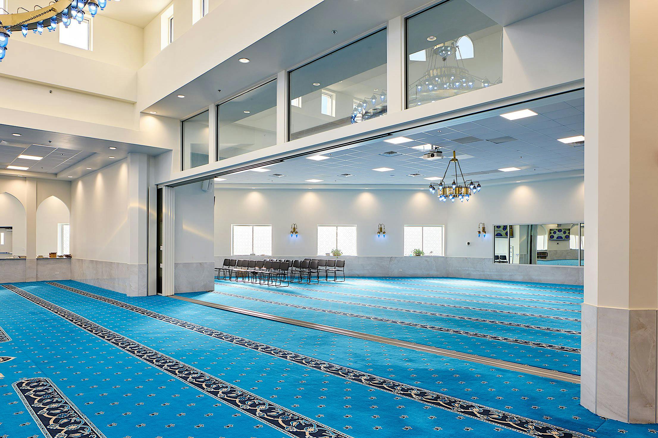 The interior of a mosque features blue carpets, white walls. Large sliding glass doors invite ample natural light, enhancing the room's bright and airy feel.