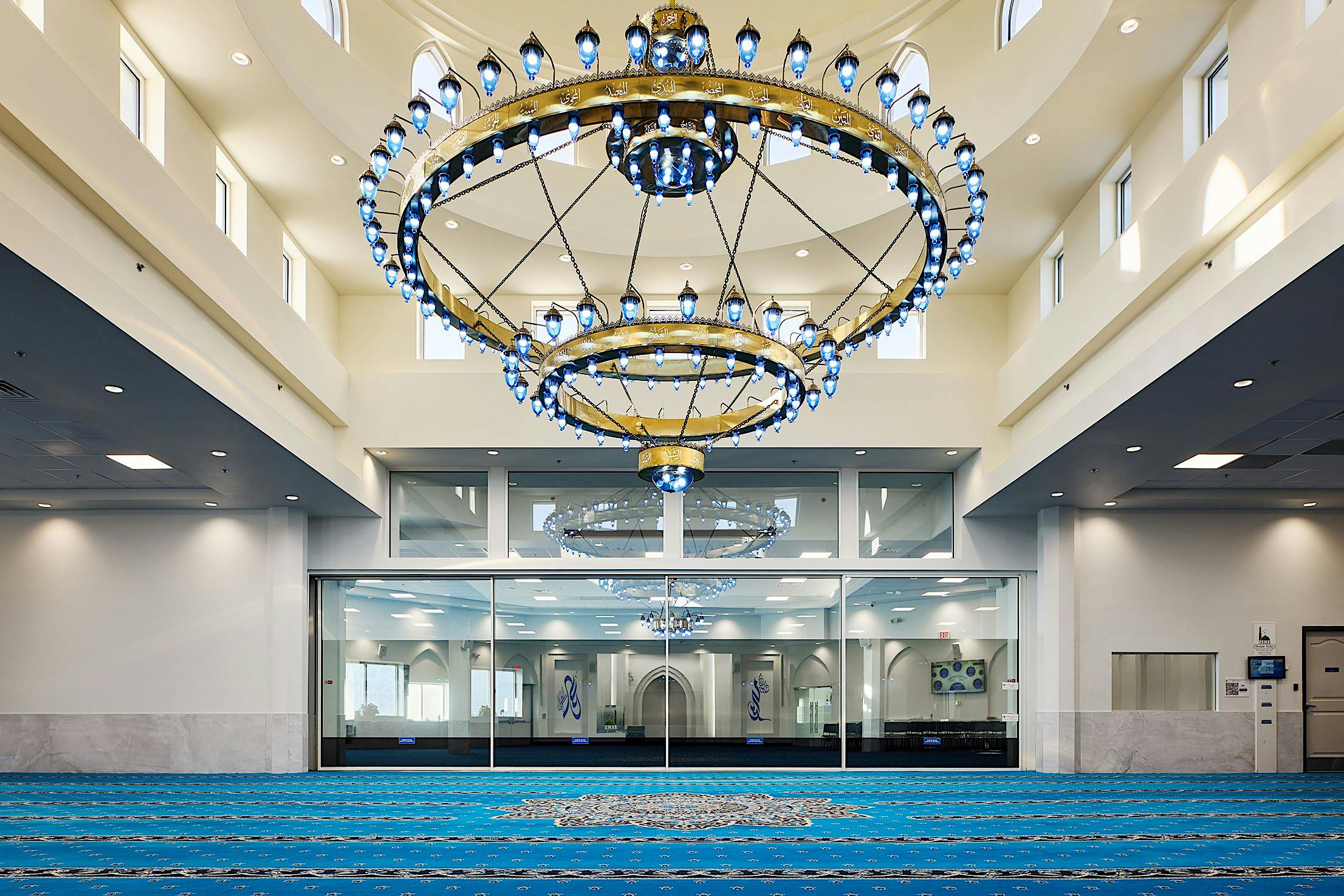 large, ornate chandelier hangs in a spacious mosque interior with blue carpet and white walls, featuring tall sliding glass doors with large transoms.