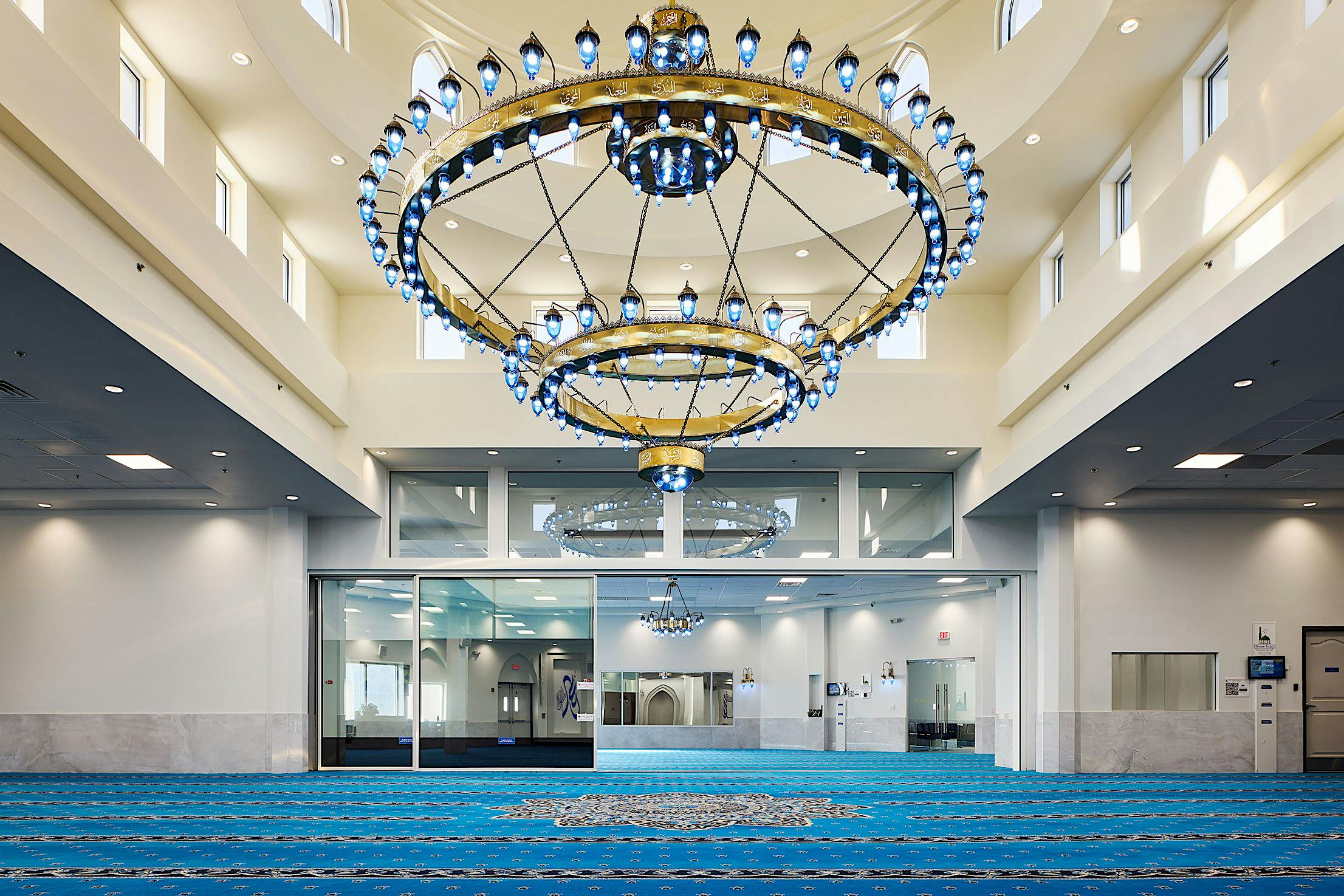 Interior of a mosque with large blue carpet, a grand chandelier, and a glass partition dividing the space.