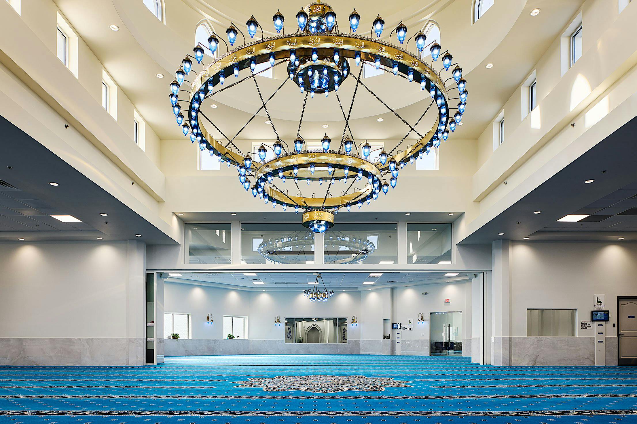 Interior of a mosque featuring a large circular chandelier, blue carpet with geometric patterns, and high ceilings with large sliding glass doors.