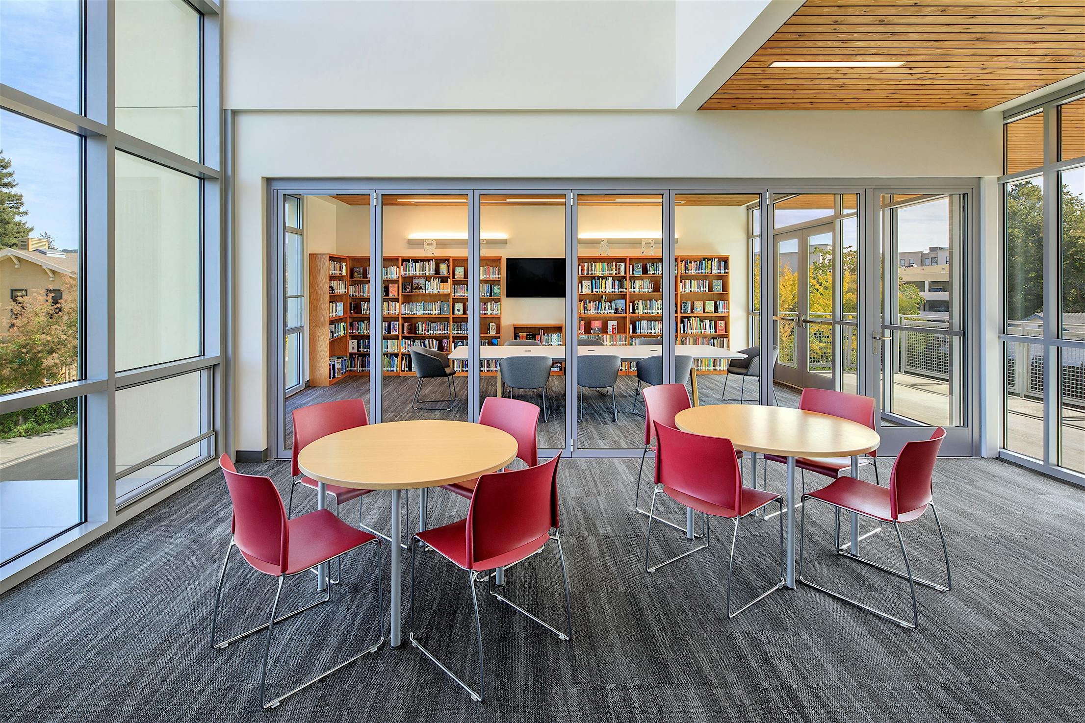 Modern library interior with large accordion glass doors, bookshelves, a round table with red chairs, and wood-paneled ceiling.