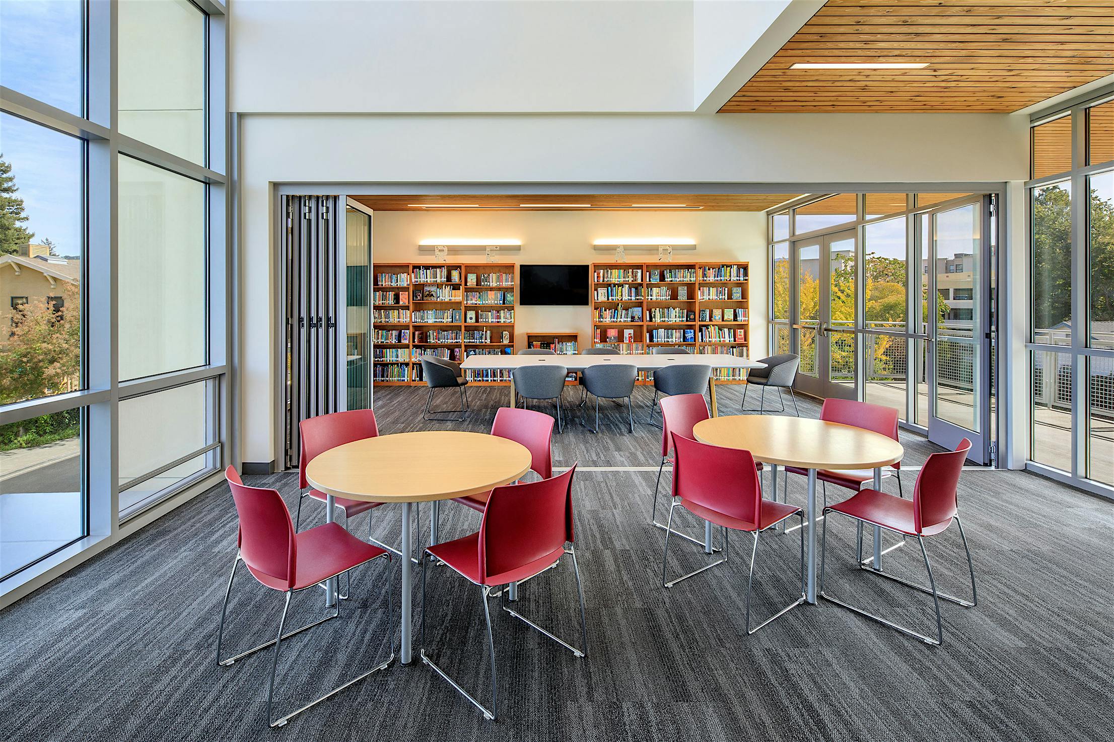 Modern library interior with opening glass doors, large windows, round tables with red chairs, and bookshelves.