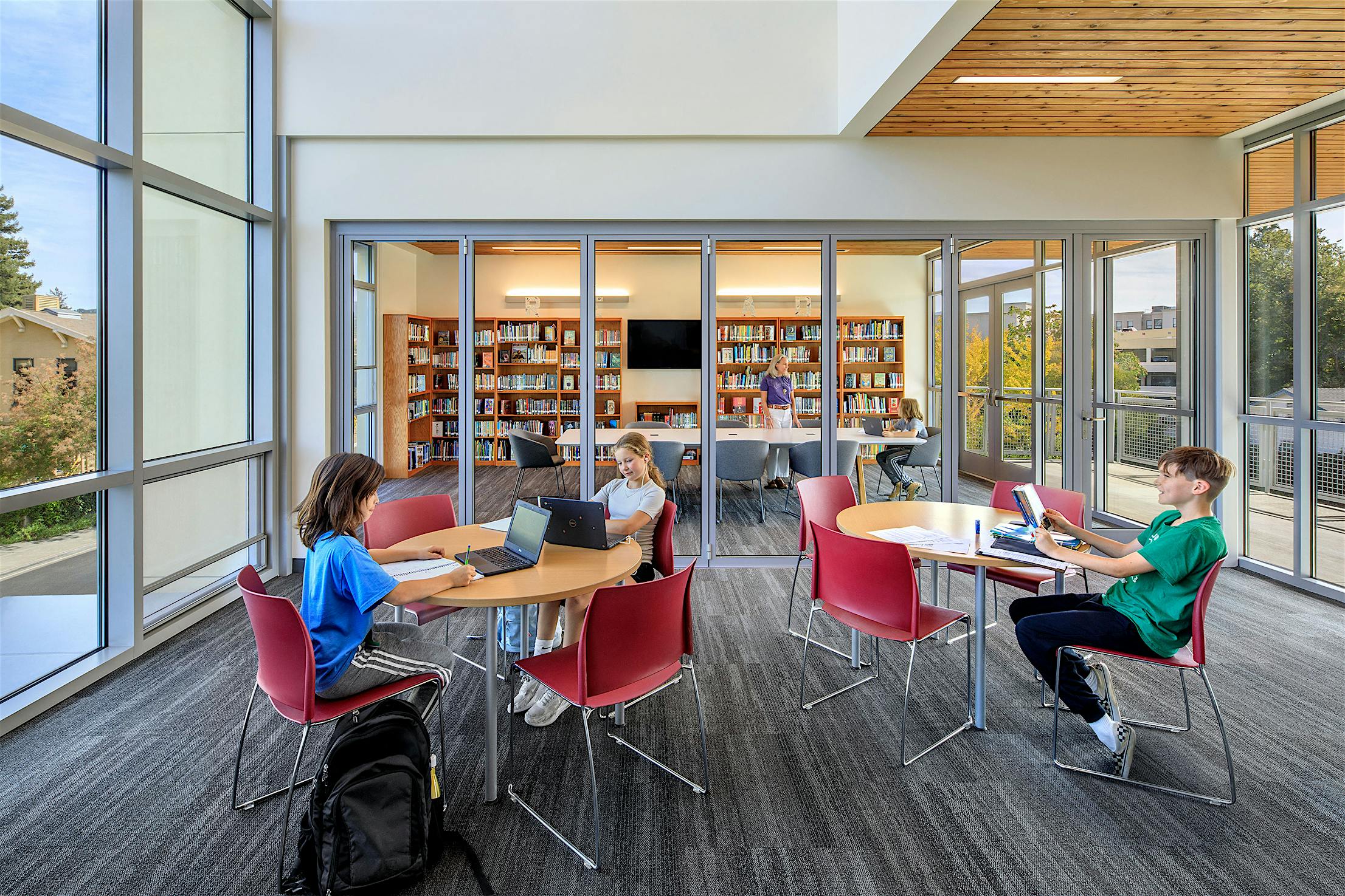 Modern library interior with opening glass doors, large windows, round tables with red chairs, and bookshelves.
