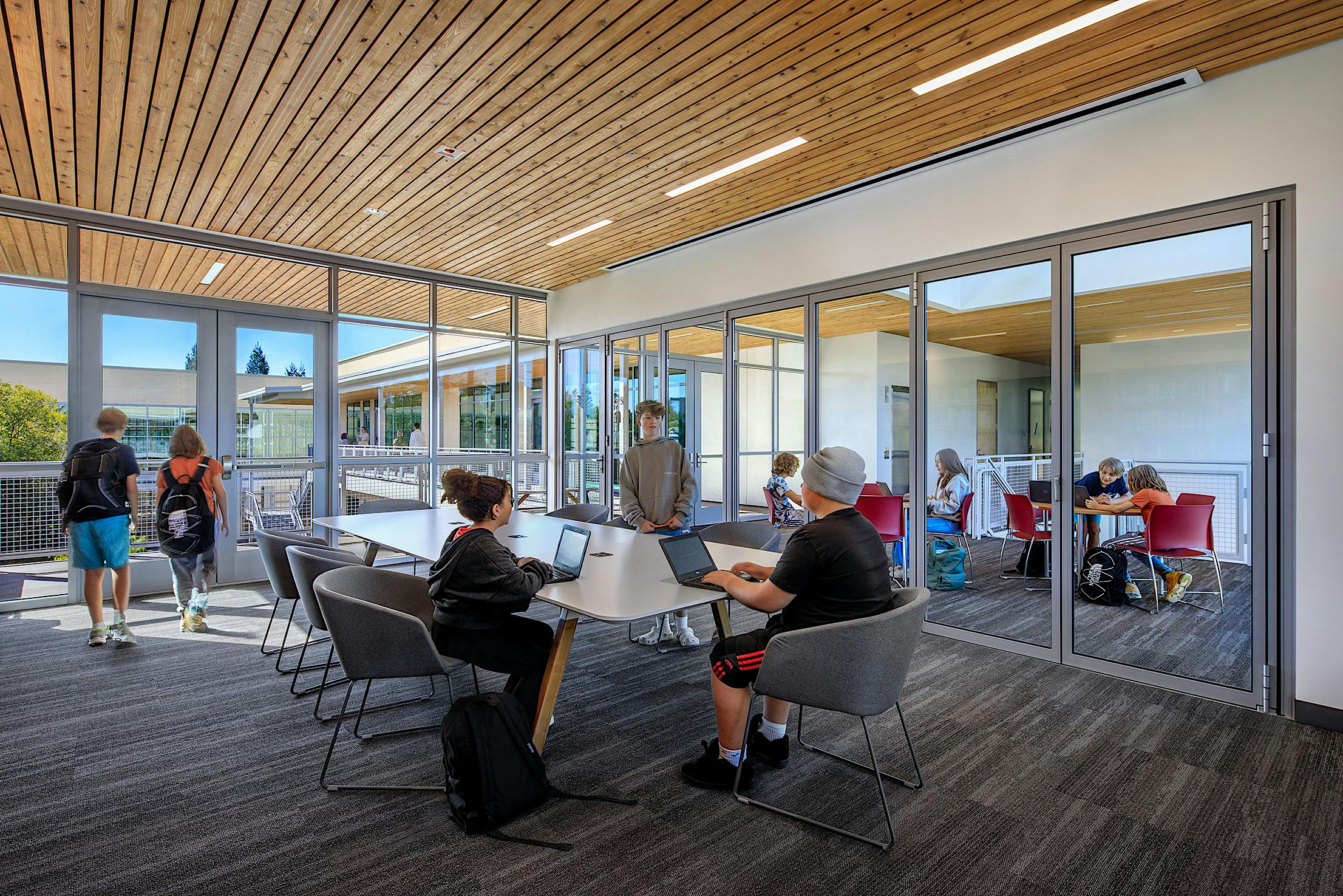Students in a modern study area with laptops and near a glass-walled room.
