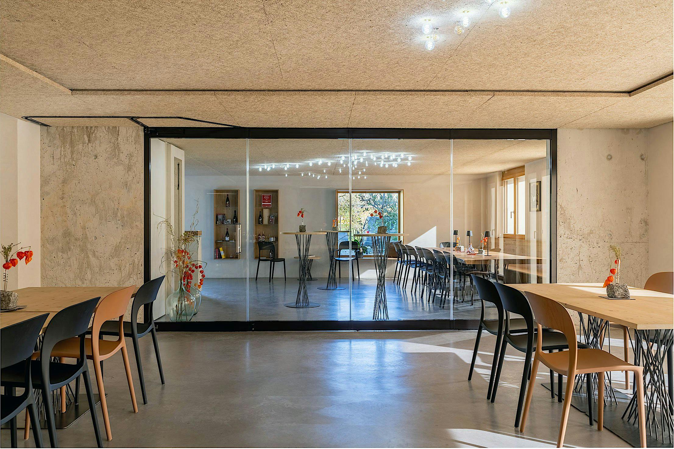 A modern wine tasting area with wooden tables and mixed black and brown chairs features sliding glass doors as a partition, seamlessly separating it from another room with similar seating.