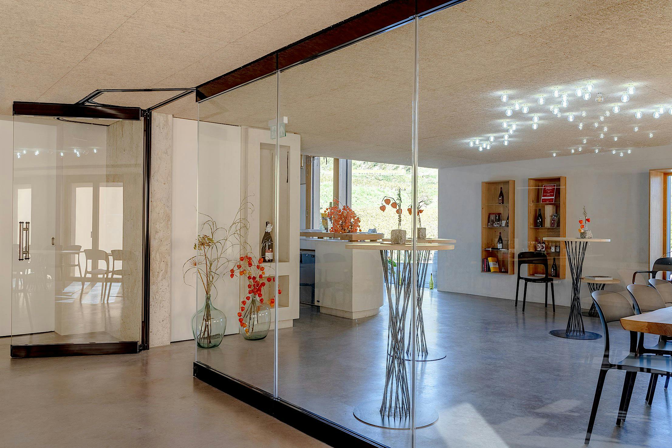 Modern tasting room interior with glass walls, high tables with stools, decorative vases with flowers, and ceiling lights.