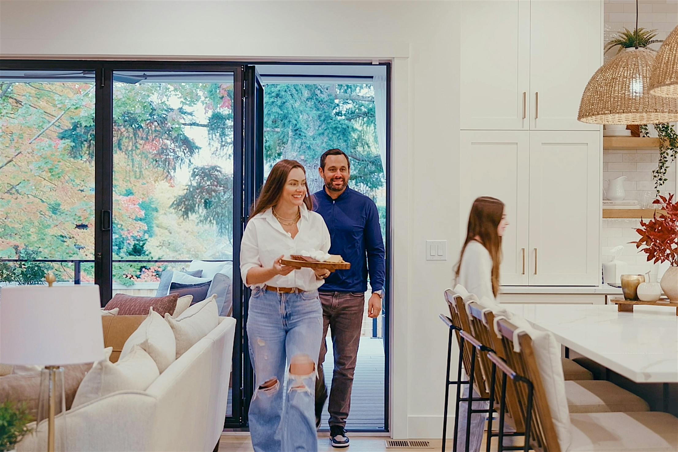 Molly Mesnick carrying a dish and a man enter a kitchen through the bifold glass doors, trees sway gently outside.