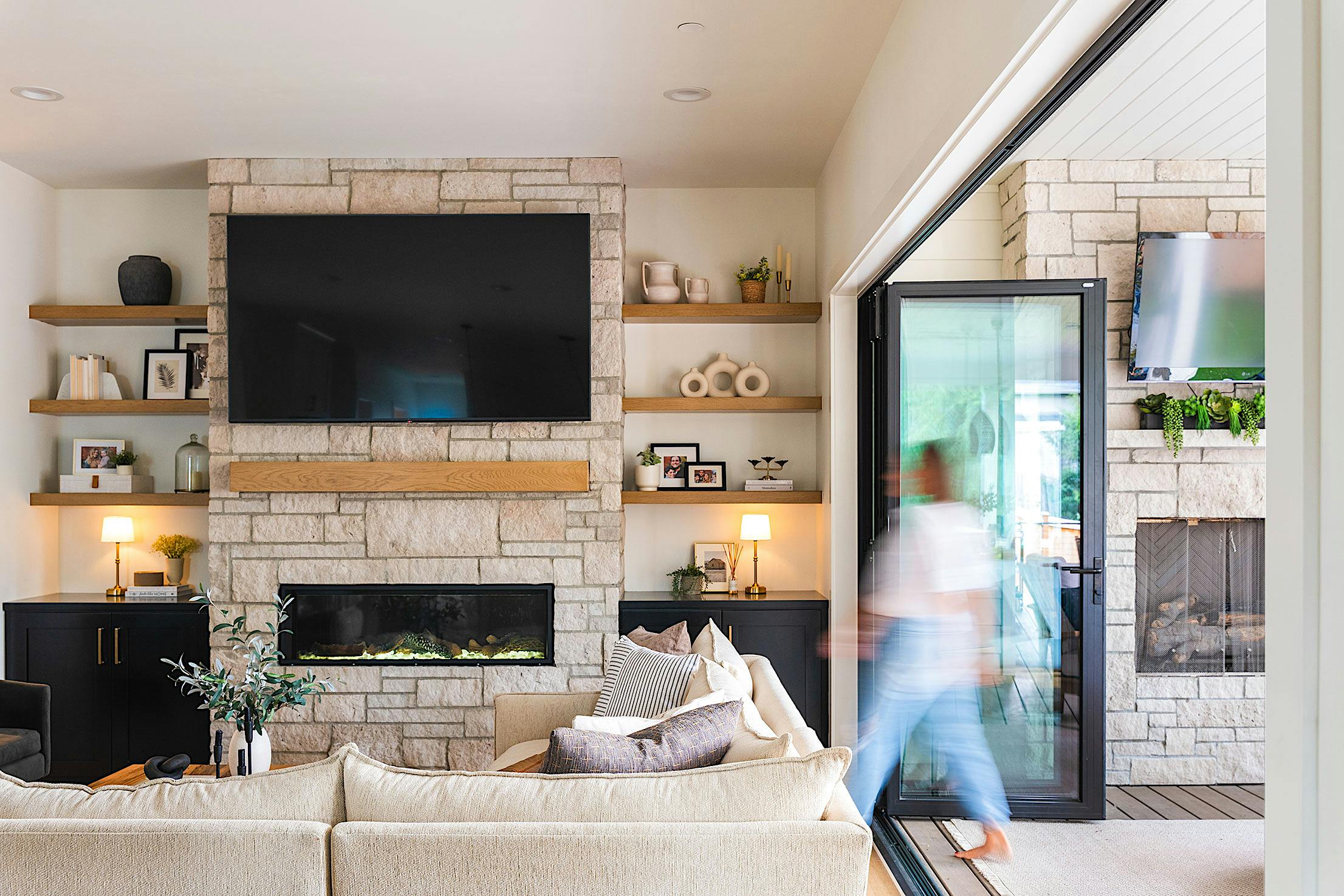 Molly Mesnick is walking past sleek bifold glass doors leading to an outdoor area with another fireplace.