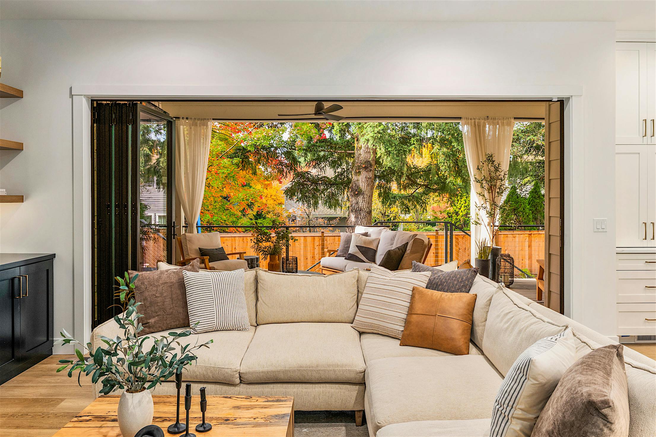The living room, featuring a beige sectional sofa and a wooden coffee table, is enhanced by bifold glass doors that open to a patio with outdoor seating.