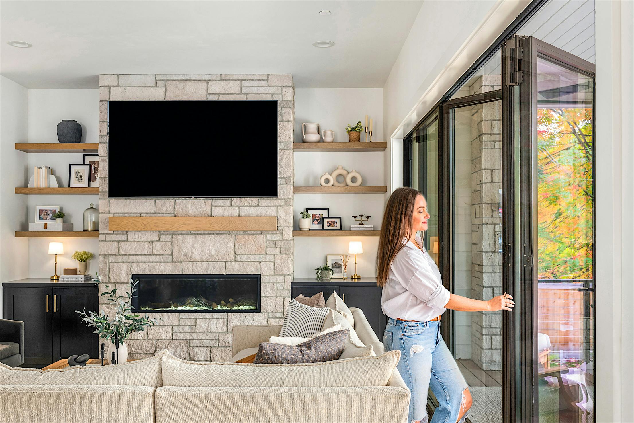 Molly Mesnick swings open the sleek bifold glass doors in a modern living room, revealing a stone fireplace, wall-mounted TV, and beige sofa.