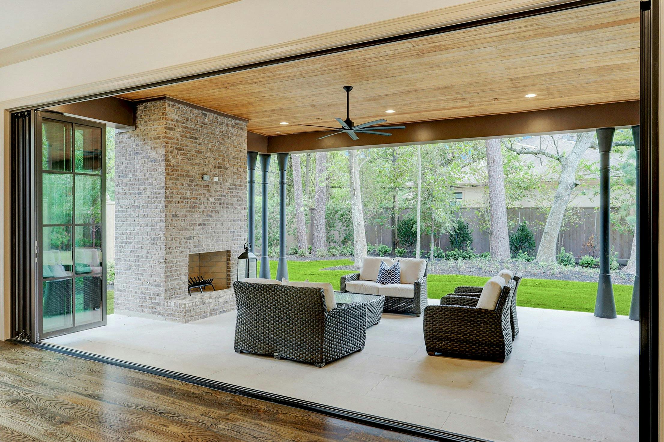 Covered patio with wicker seating, brick fireplace, and open bifold doors leading to a lush, tree-lined backyard.