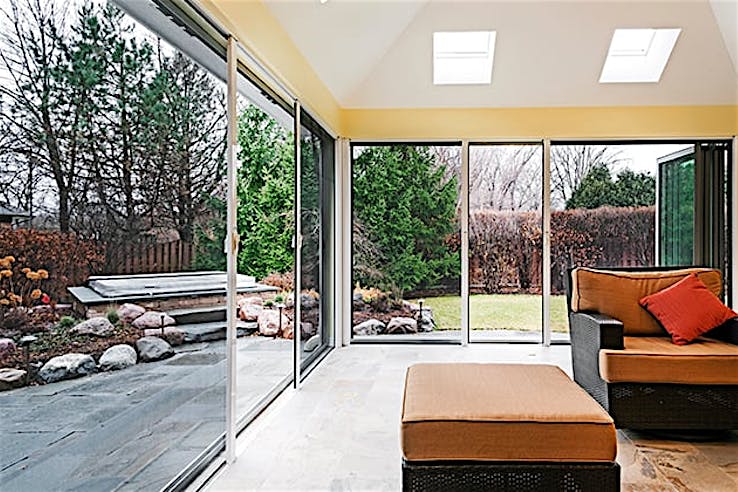 Sunroom with sliding glass doors and screens opens to a landscaped backyard with trees.
