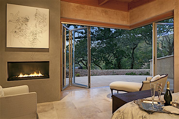 Modern living room with a fireplace, abstract wall art, and open corner accordion glass doors leading to a patio with a view of trees.