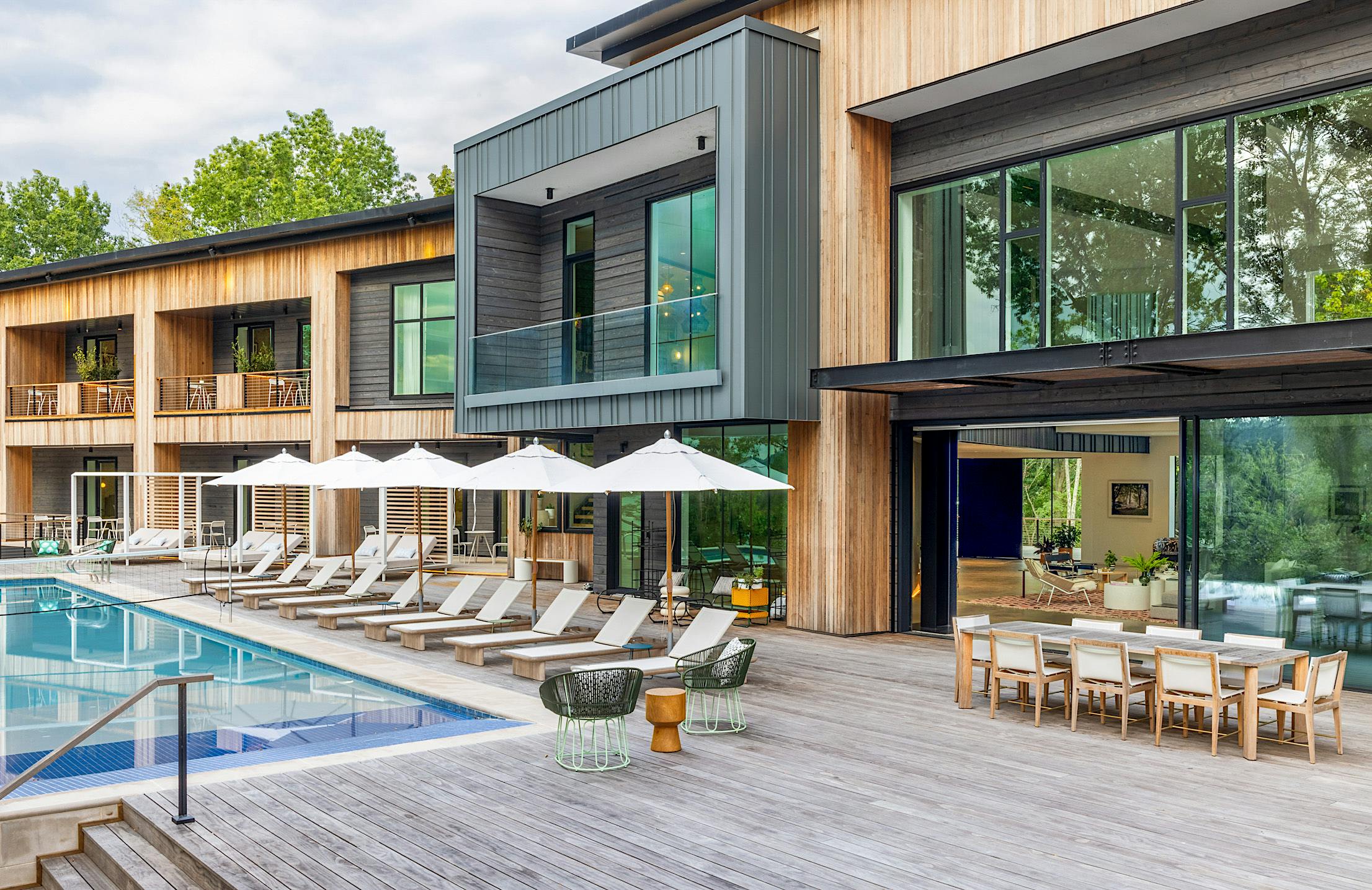 Modern house with large opening glass doors, outdoor swimming pool, lounge chairs with umbrellas, and spacious deck with dining area.