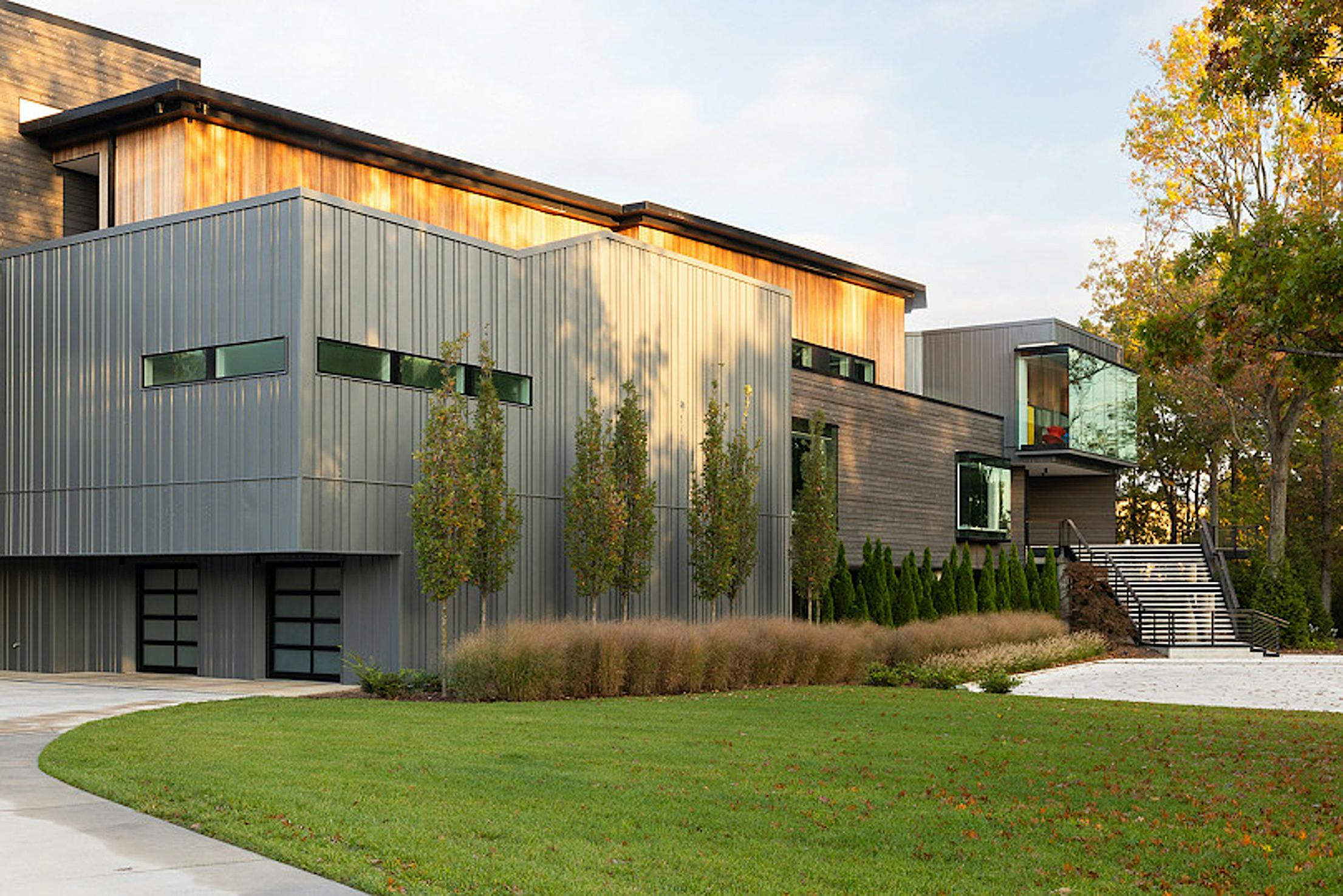 Modern two-story home with metal and wood exterior, large transom windows, and landscaped front yard, daytime driveway view.