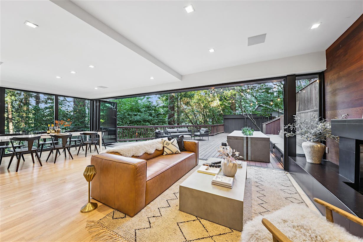 Patio glass doors open from a modern living area to a spacious outdoor deck surrounded by trees.