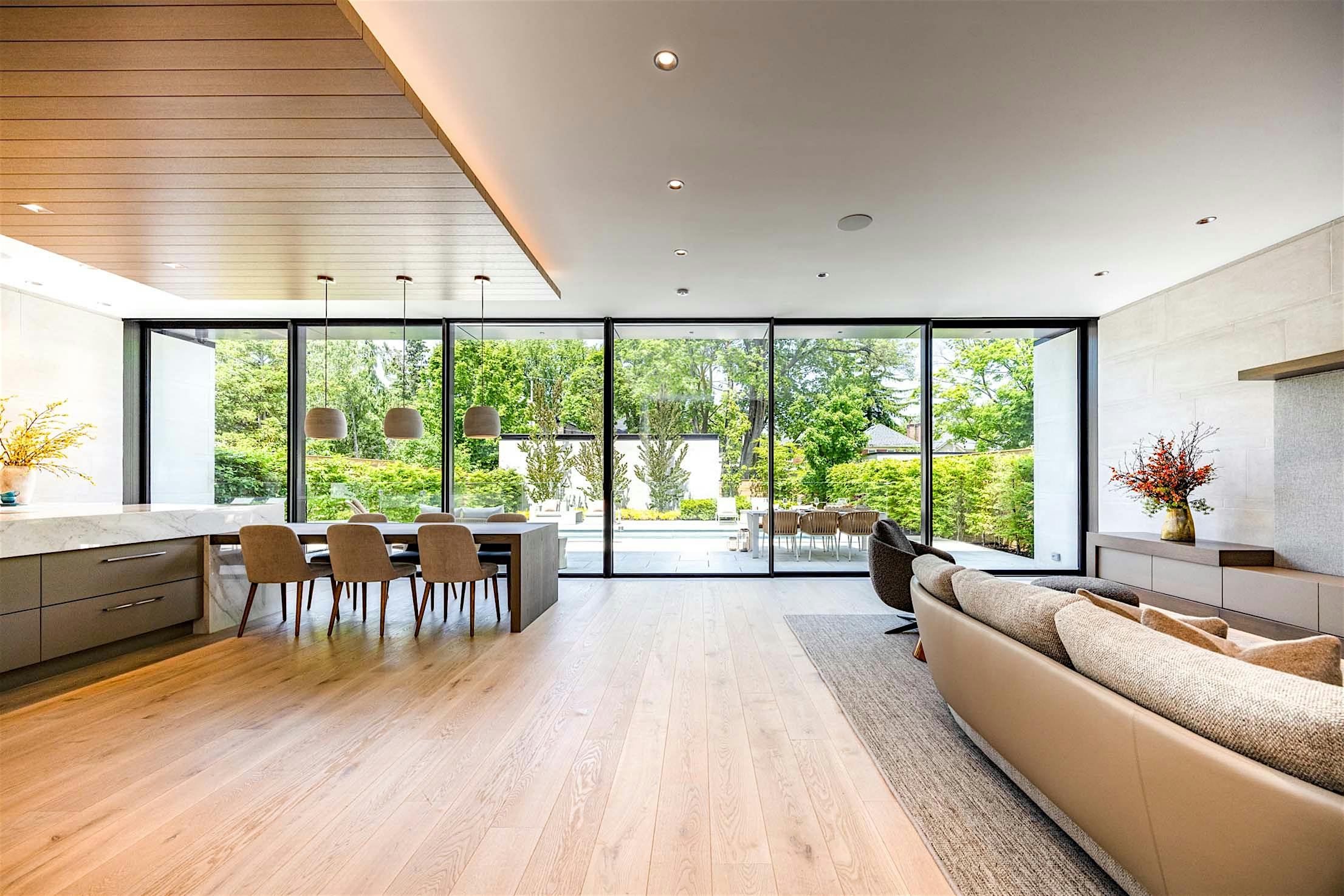 Large sliding glass doors reveal a modern open-plan living and dining area with neutral furniture, wood flooring, and garden views